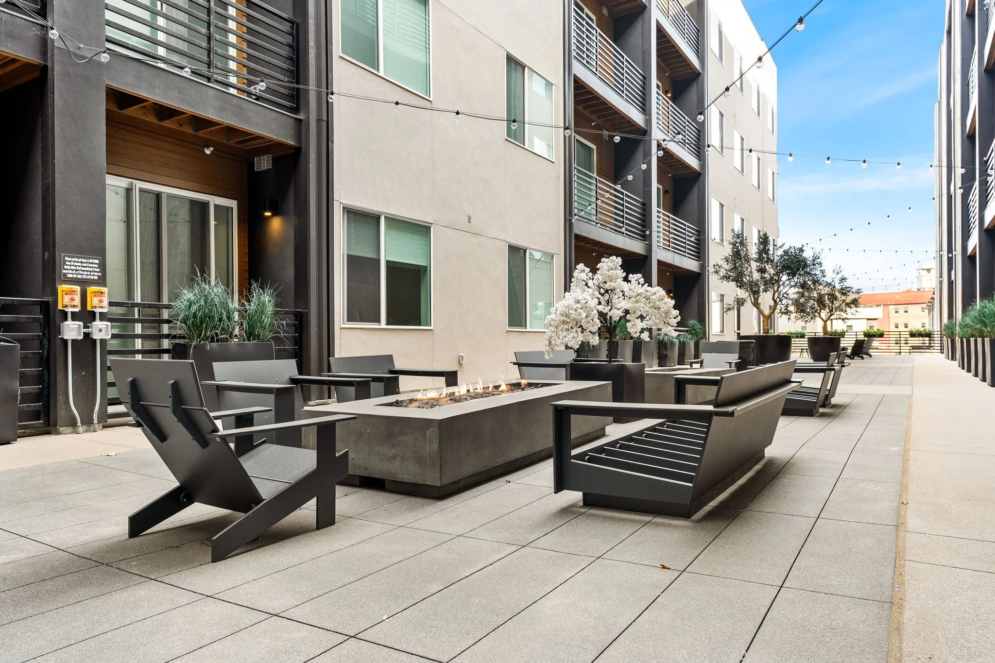 Outdoor patio with modern black benches and planters with white flowers and small trees, lined with string lights, in front of a multi-story residential building with beige and dark gray walls.