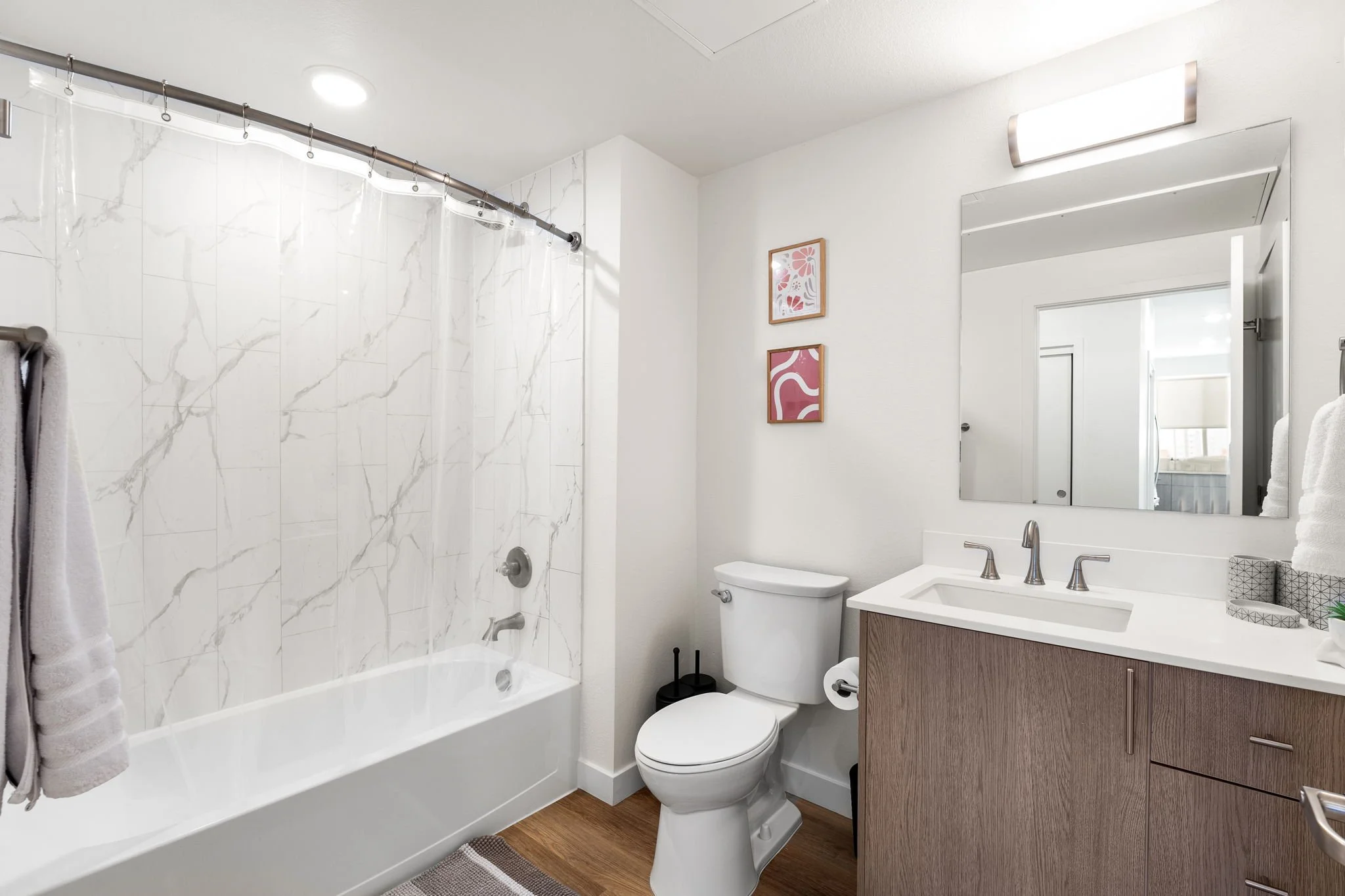 Modern bathroom with a bathtub and shower, a toilet, and a wooden vanity with a sink, mirror, towel, and artwork on the wall.