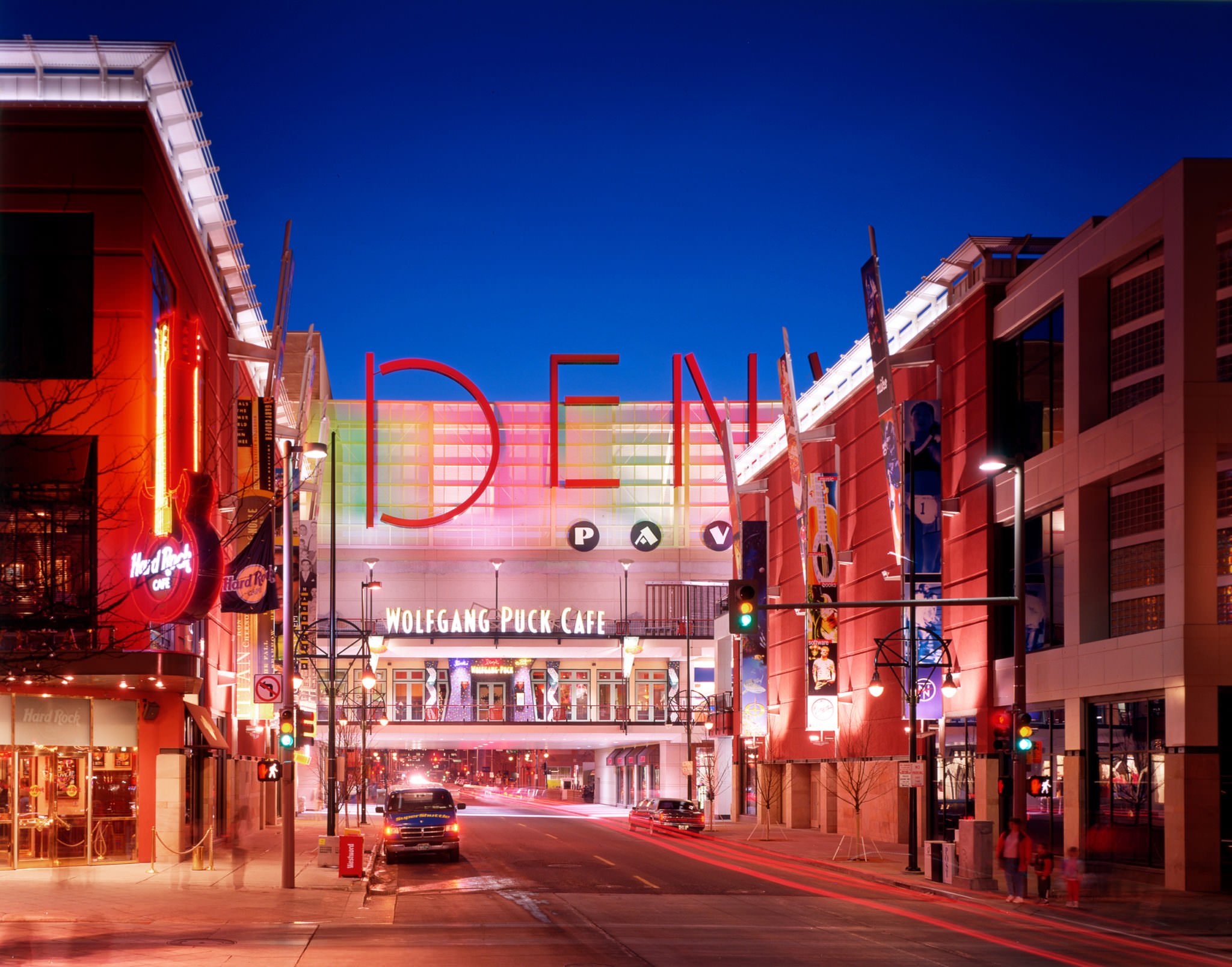 Night view of The Dome shopping mall with neon signs, including a Hard Rock Cafe guitar, and the Wolfgang Puck Cafe on the second floor. Streets are illuminated with streetlights, and there are a few cars and pedestrians.