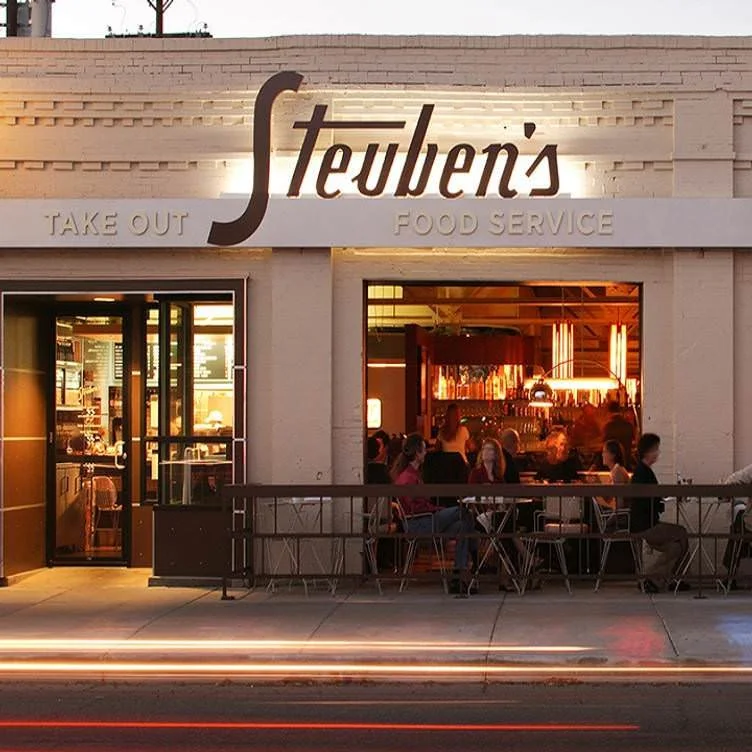 Exterior view of a restaurant called 'Steuben's,' with seating outside. The restaurant offers takeout and food service, and the interior is visible through large windows, showing people dining inside.