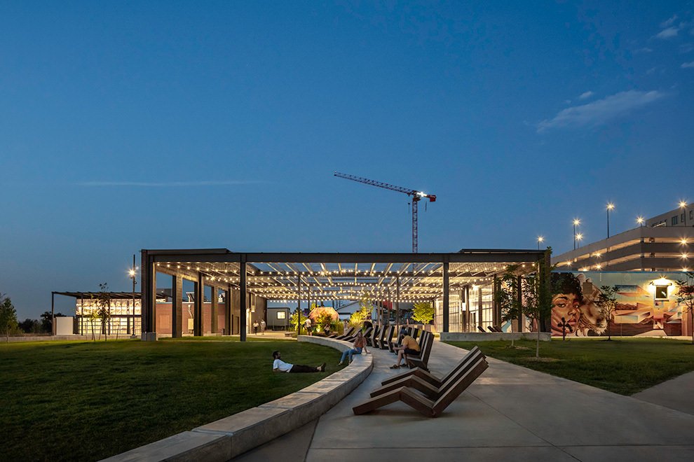 A modern outdoor space at dusk with a lit pavilion, benches, grass, and a colorful mural on a building wall.