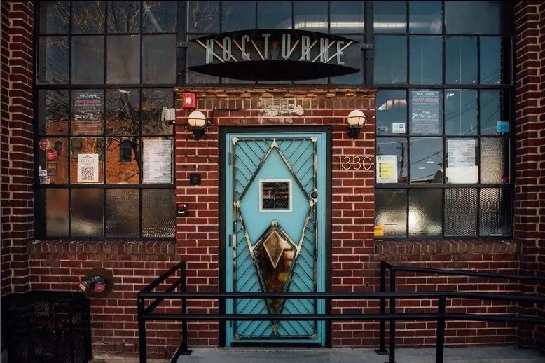 Exterior of a brick building with large industrial-style windows, a teal decorative door with geometric patterns, two wall-mounted lights, a black awning with the words 'NOCTURNE,' and a small safety sign on the door.
