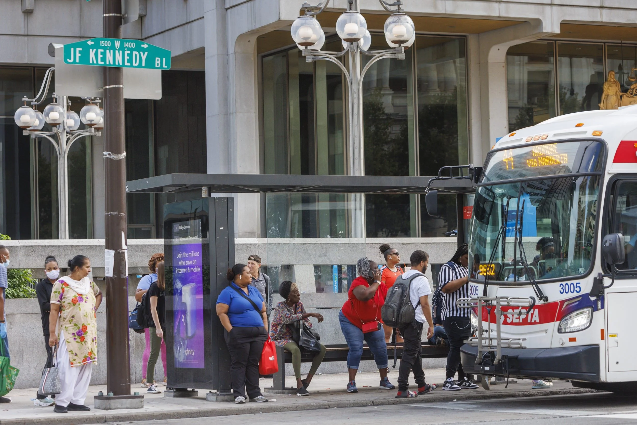 People boarding a SEPTA bus at a city bus stop on JF Kennedy Boulevard with modern buildings and street signs in the background.