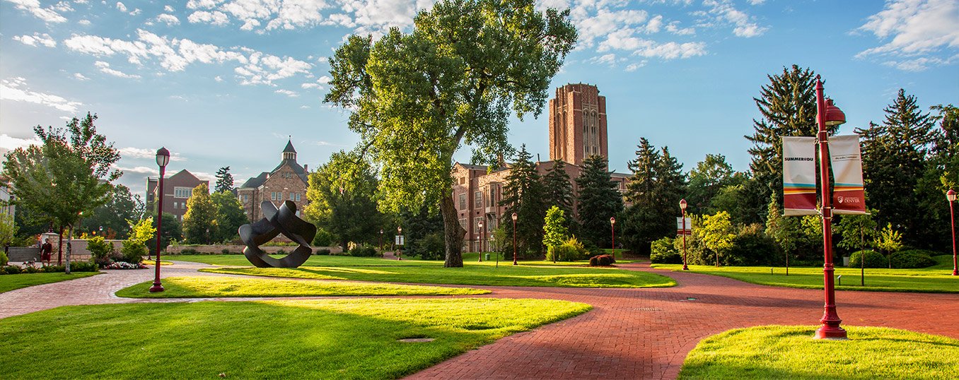 A well-maintained park on a bright, sunny day with a paved red brick pathway, lush green grass, mature trees, a modern sculpture, and historic buildings in the background surrounding the University of Denver.