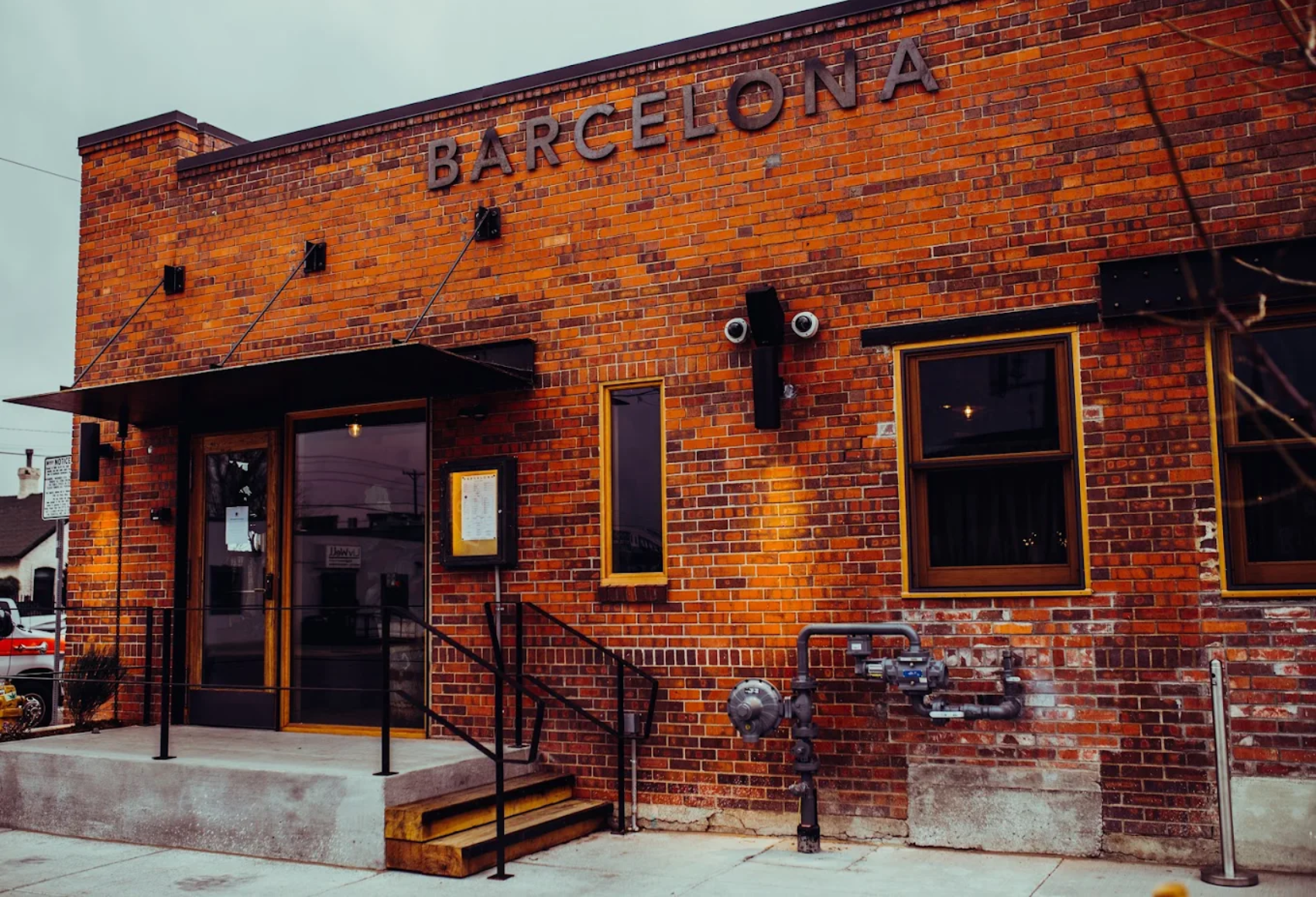 Exterior view of a brick restaurant with the name 'BarceloNa' on the wall, featuring a small concrete entry porch with steps, black metal handrails, and windows with brown frames. Security cameras and piping are visible on the brick wall.