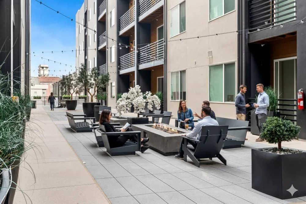 An outdoor courtyard enclosed by modern multi-story apartment buildings with string lights hanging overhead. There are planters with green plants and benches along the walkway, with larger potted plants in the foreground and a tree with white flowers in the center.