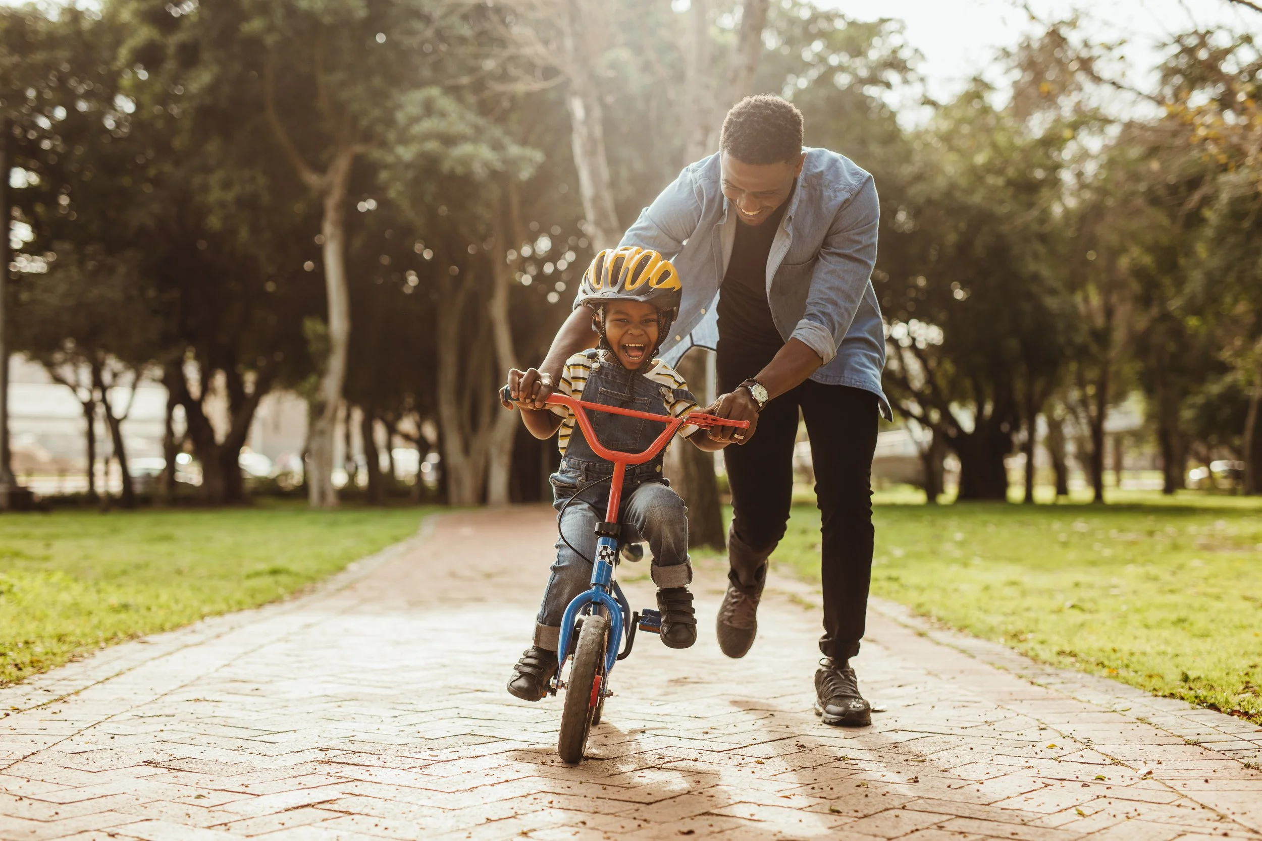 A young boy learning to ride a bicycle with training wheels while his father helps him, both smiling, in a park with trees and grass in the background.