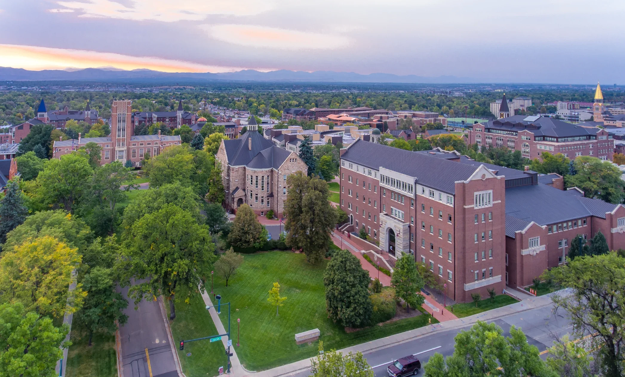 An aerial view of a university campus with historic and modern brick buildings, green trees, sidewalks, and a street with cars at sunset, with mountains in the background.