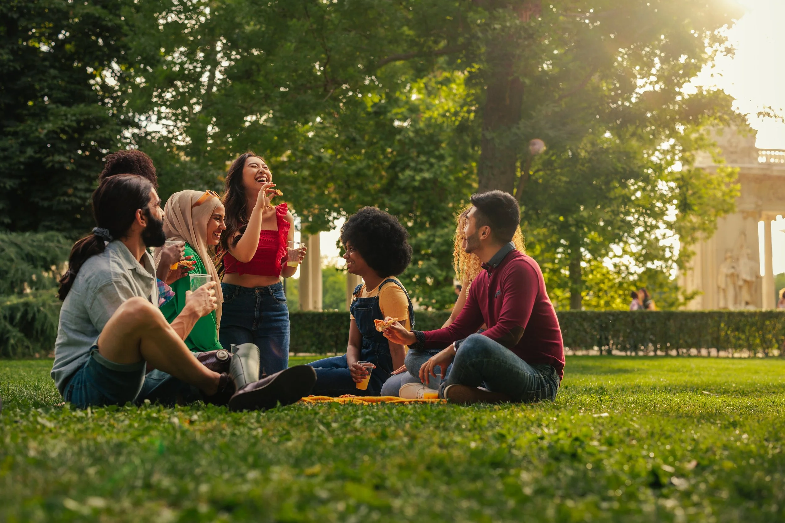 Group of friends having a picnic in a park on a sunny day, sitting on the grass, talking, laughing, and holding drinks and snacks, with trees and a historic building in the background.