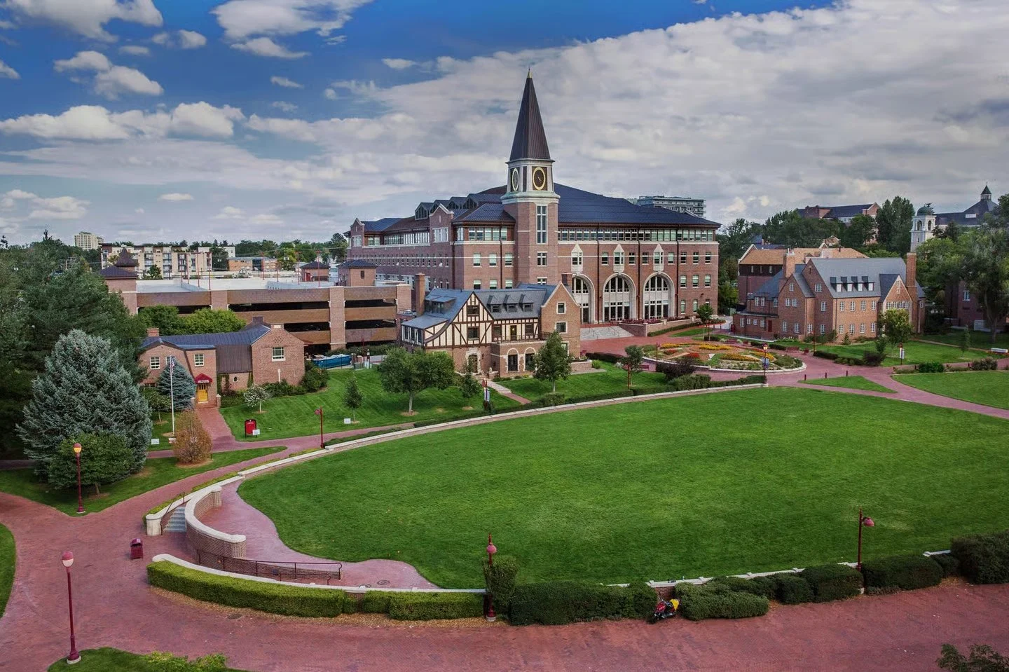 View of a university campus with a large brick building featuring a clock tower, surrounded by smaller brick and Tudor-style buildings, a green lawn, paved pathways, and trees under a partly cloudy sky.