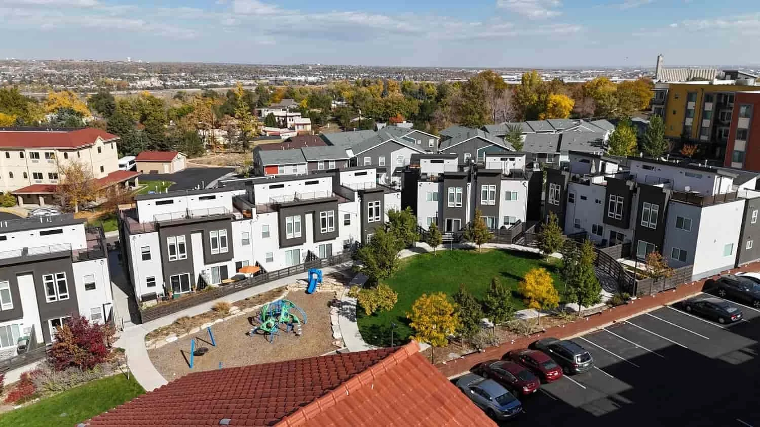 An aerial view of modern white and gray townhouses with rooftop decks, a small playground with swings and a slide, a green lawn with trees, and a parking lot with several cars