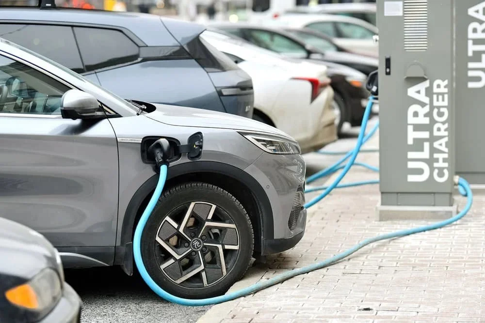 A person in a gray sweater and yellow vest interacts with a smartphone, with a black electric vehicle charger attached to a brick wall in the background.