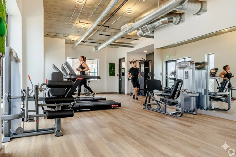 Modern gym with cardio machines, strength training equipment, and wooden flooring, large windows, and exposed ductwork on the ceiling.