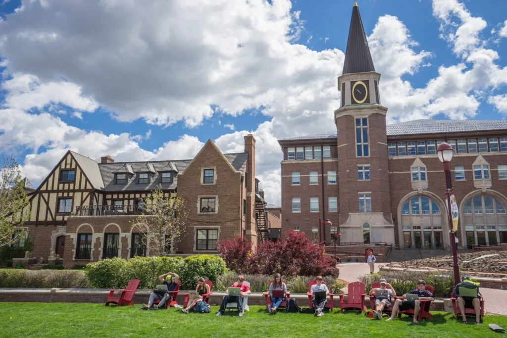 People sitting on red chairs in a park, using laptops and phones, with a building featuring a clock tower in the background and a partly cloudy sky.