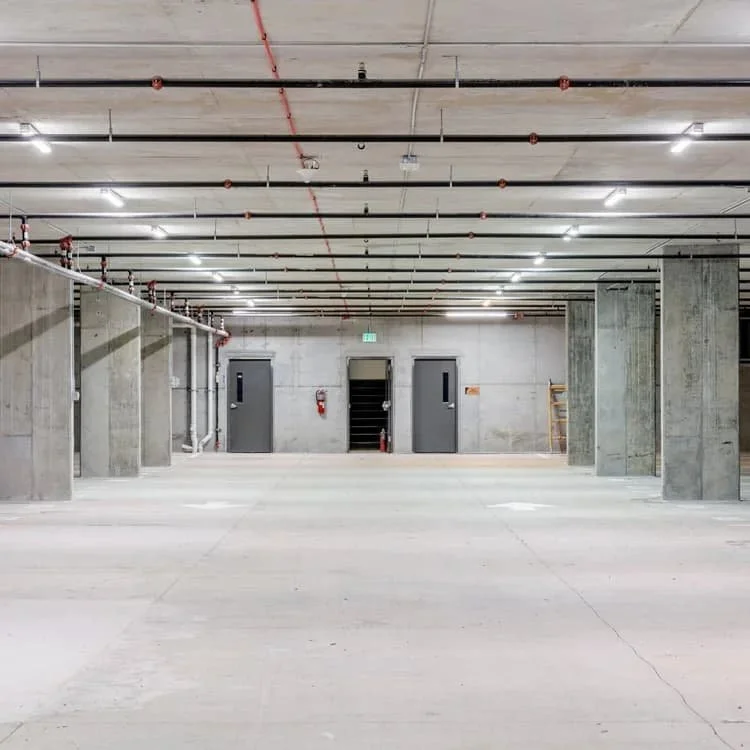Underground parking garage with concrete walls, columns, ceilings, and floors, featuring fire sprinklers, emergency exit signs, and fire extinguishers.