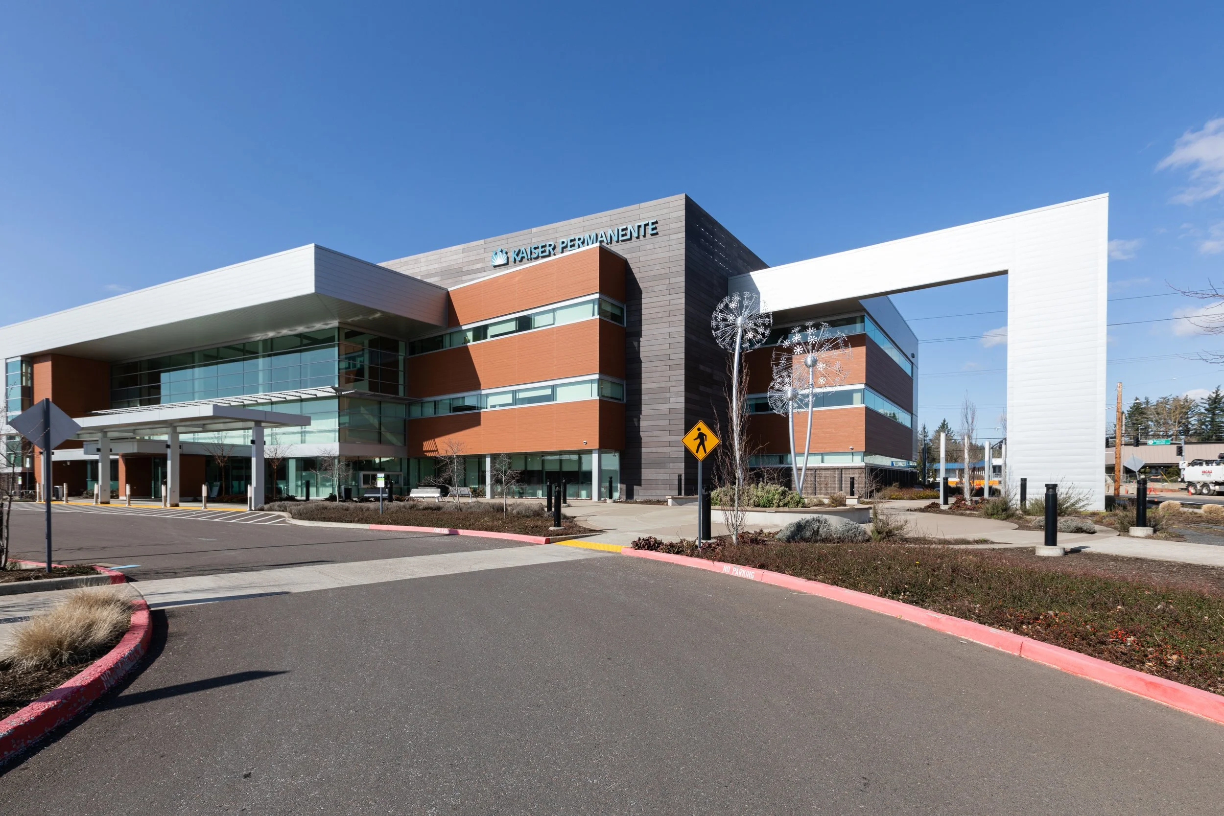 Modern hospital building with signs that read 'Kaiser Permanente,' a parking lot with no parked cars, decorative metal dandelion sculptures, and a pedestrian crossing sign, under a clear blue sky.