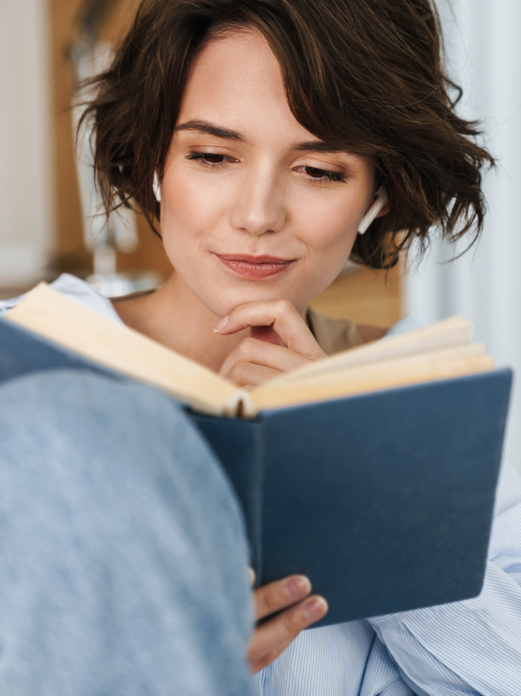A woman with short brown hair wearing wireless earbuds is holding a book and smiling softly, appearing to be reading.