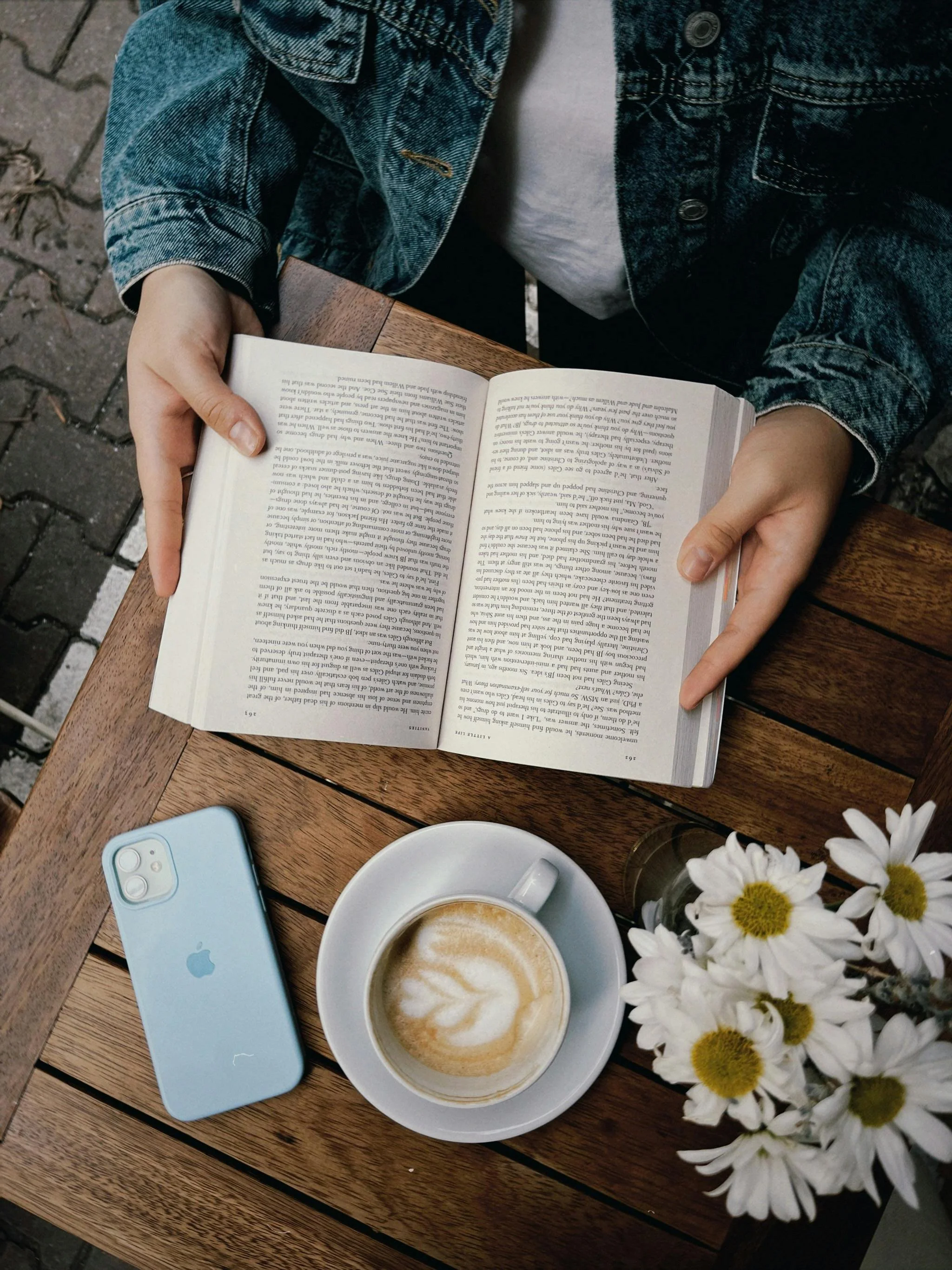 A person sitting at a wooden table with an open book, a smartphone, a cup of latte with latte art, and a small bouquet of daisies.