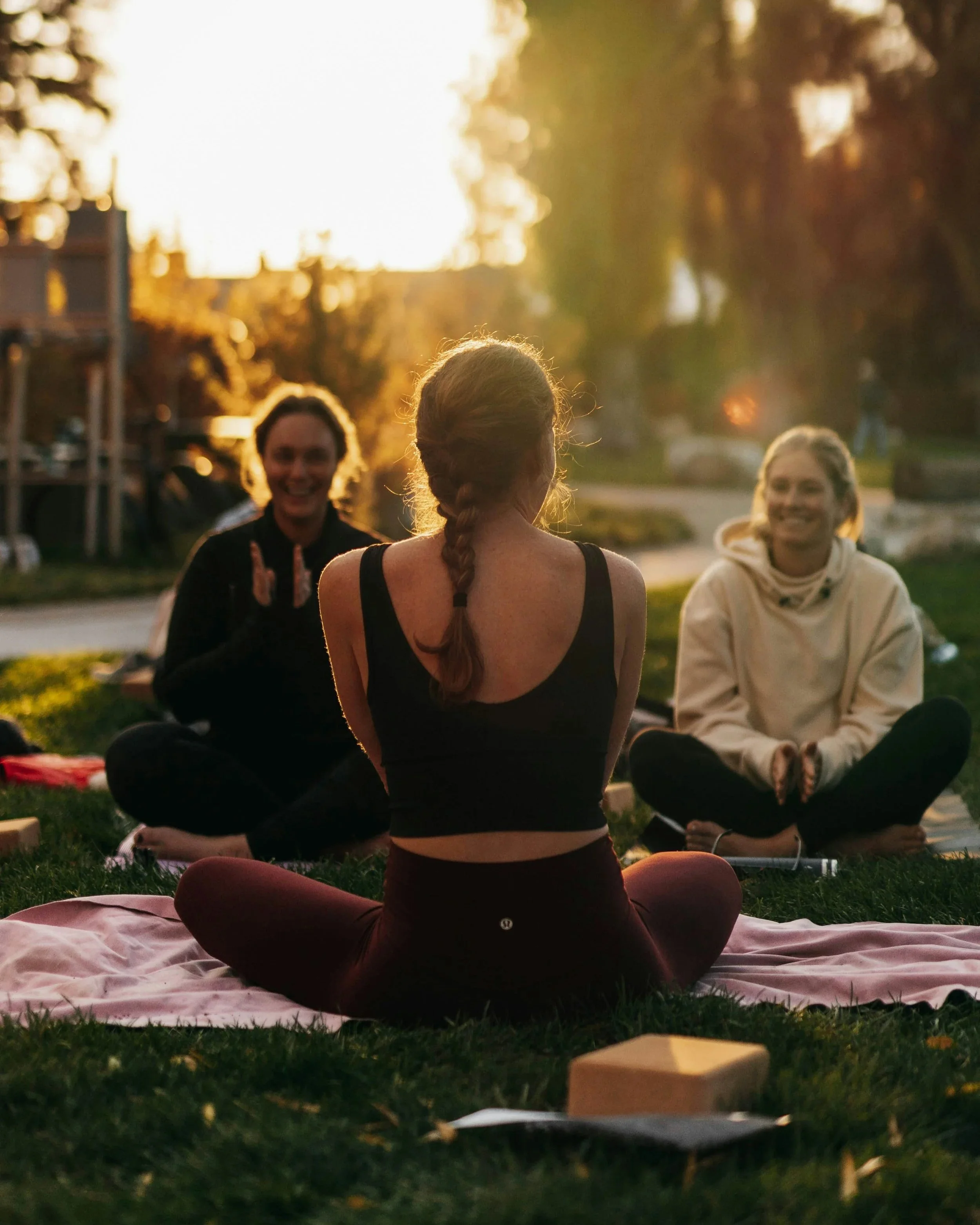 A woman seated on a blanket at a park looks across a circle at other smiling women.