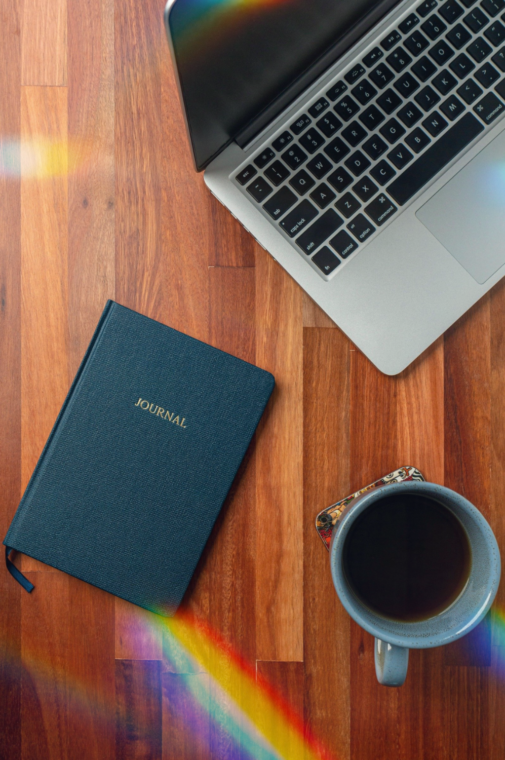 A computer, journal, and coffee cup set on a wood table shrouded in rainbows.