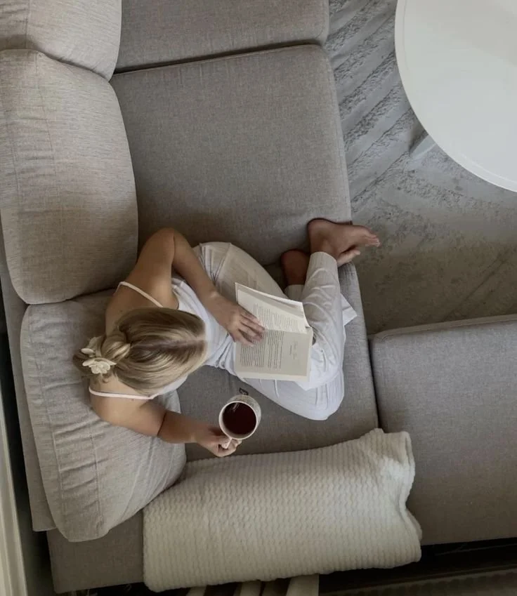 Ein junges Mädchen liegt auf einer Couch, liest ein Buch und hält eine Tasse mit einem dunklen Getränk, vermutlich Tee oder Saft, in der Hand.