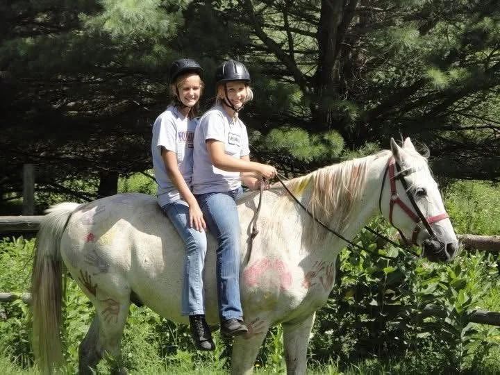 Two young girls wearing helmets riding a white horse together in a green outdoor setting.