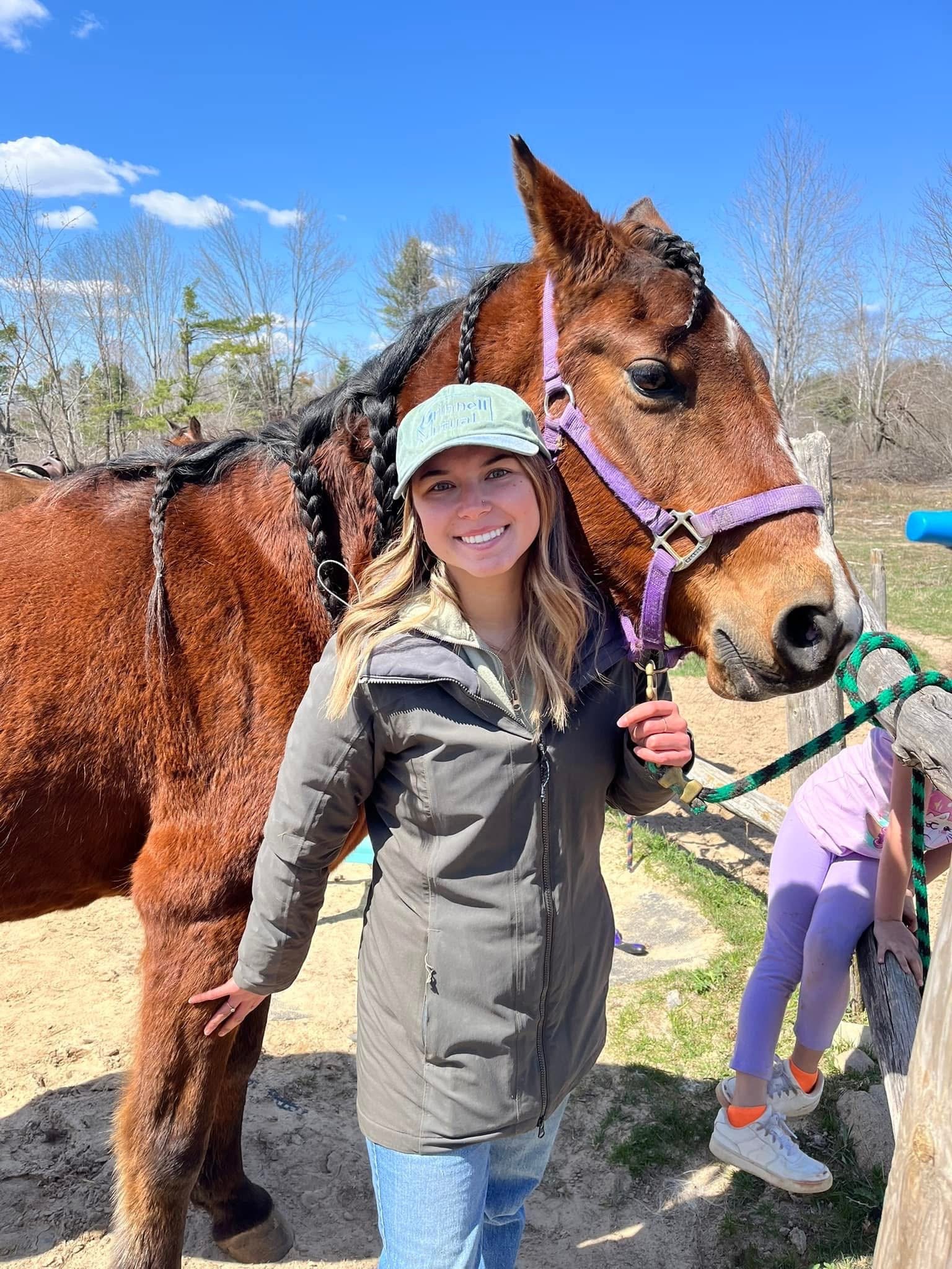 A woman with long blonde hair and a light gray hat smiling while hugging a large brown horse with a purple halter. In the background, there are other people, trees without leaves, a blue sky with scattered clouds, and a fence.