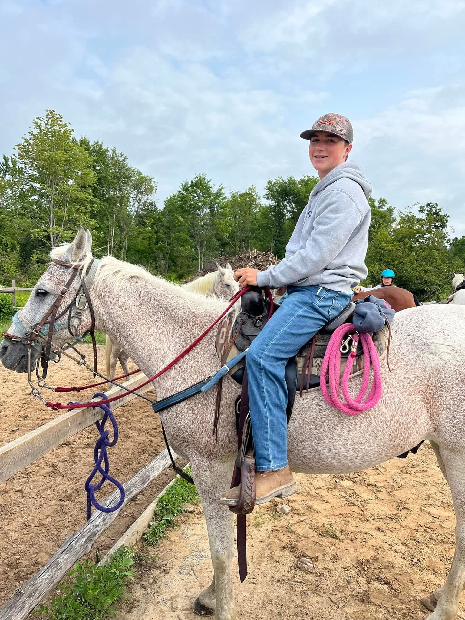 A young man in a gray hoodie and baseball cap rides a white horse with a pink rope tied to its saddle. Other riders and horses are visible in the background, set in an outdoor riding arena with a wooden fence, green trees, and a cloudy sky.