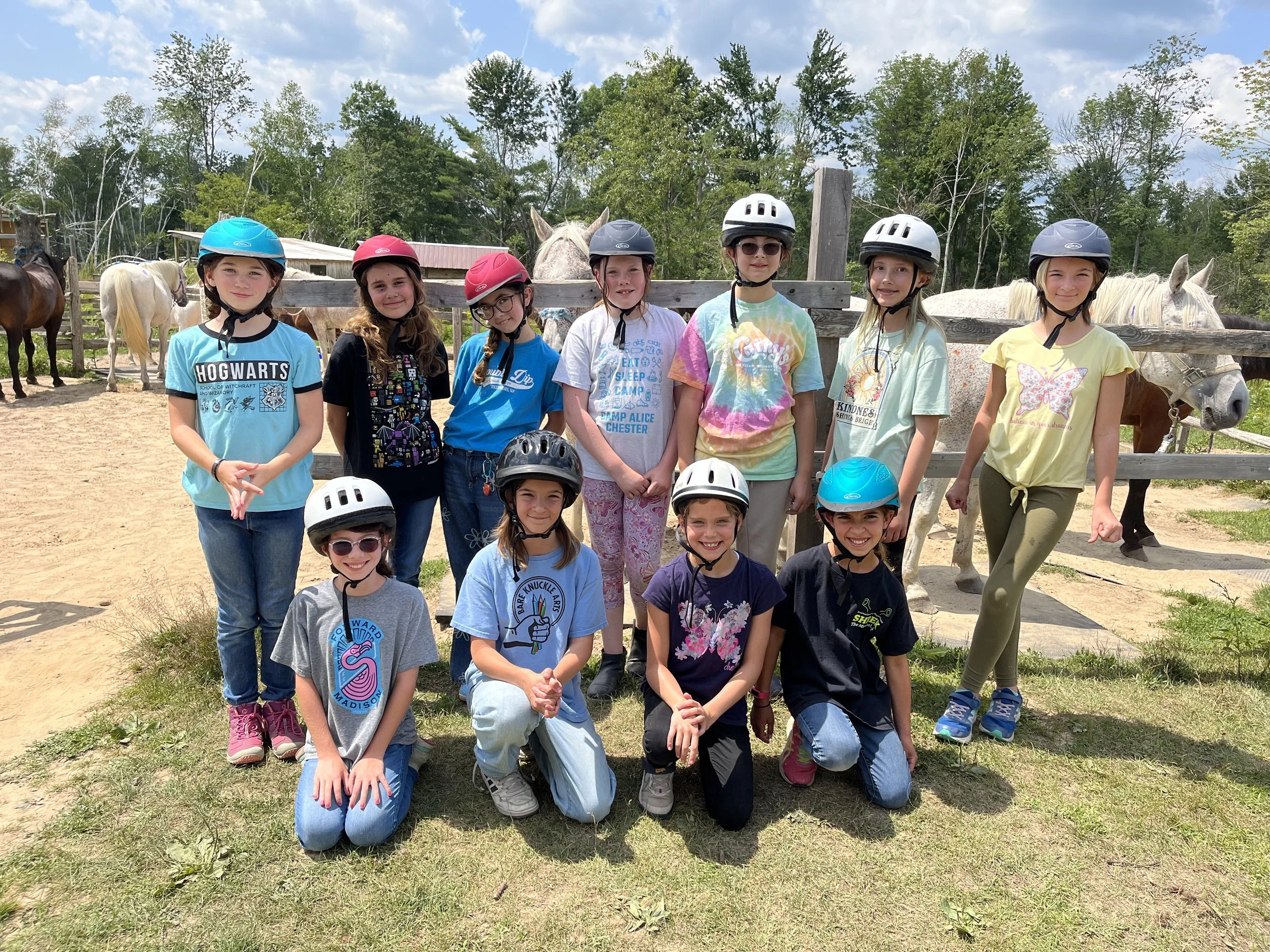 Group of children wearing helmets posing outdoors with horses behind a wooden fence and trees in the background.