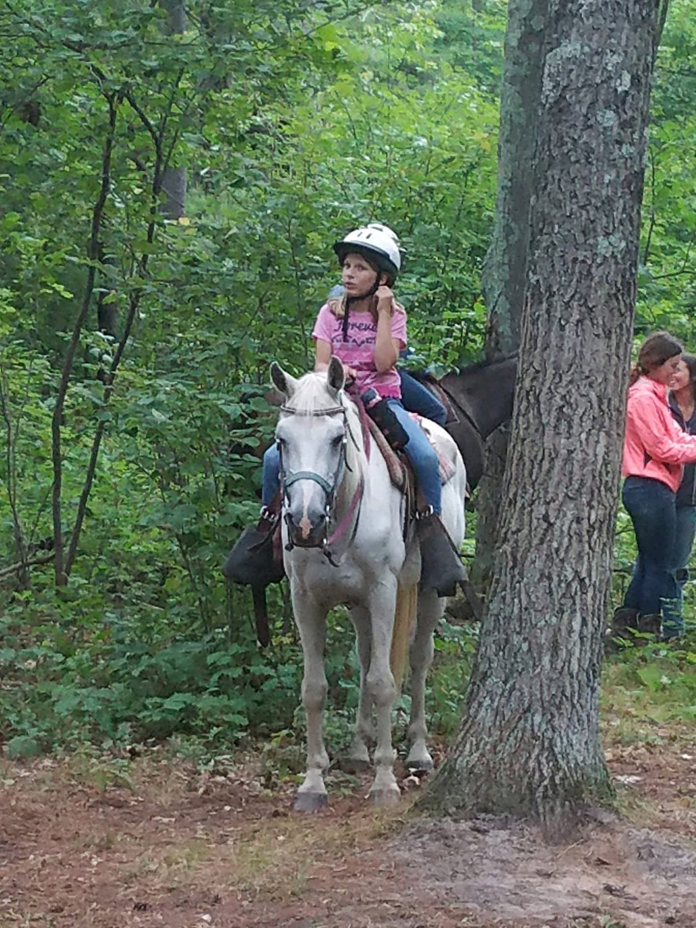 A girl riding a white horse in a wooded area with green foliage. The girl is wearing a pink shirt, blue jeans, and a white helmet. There are two women in the background, one wearing a pink jacket and the other in a darker top, standing near a tree.