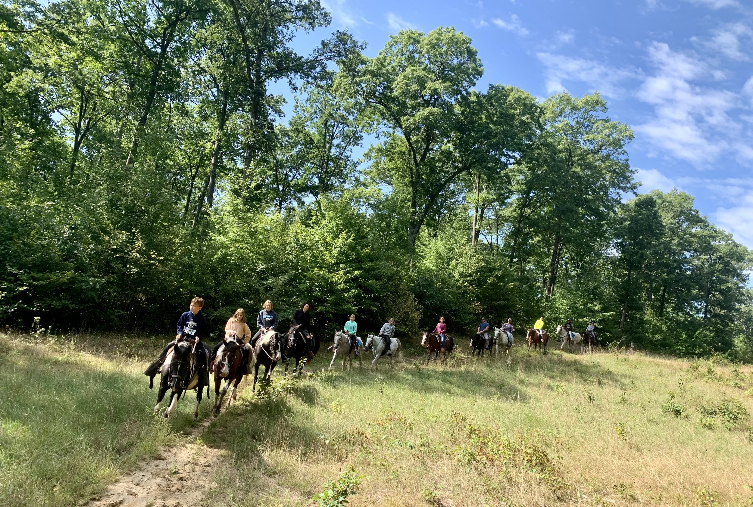 Group of children and adults riding horses in a grassy clearing surrounded by tall green trees under a partly cloudy blue sky.