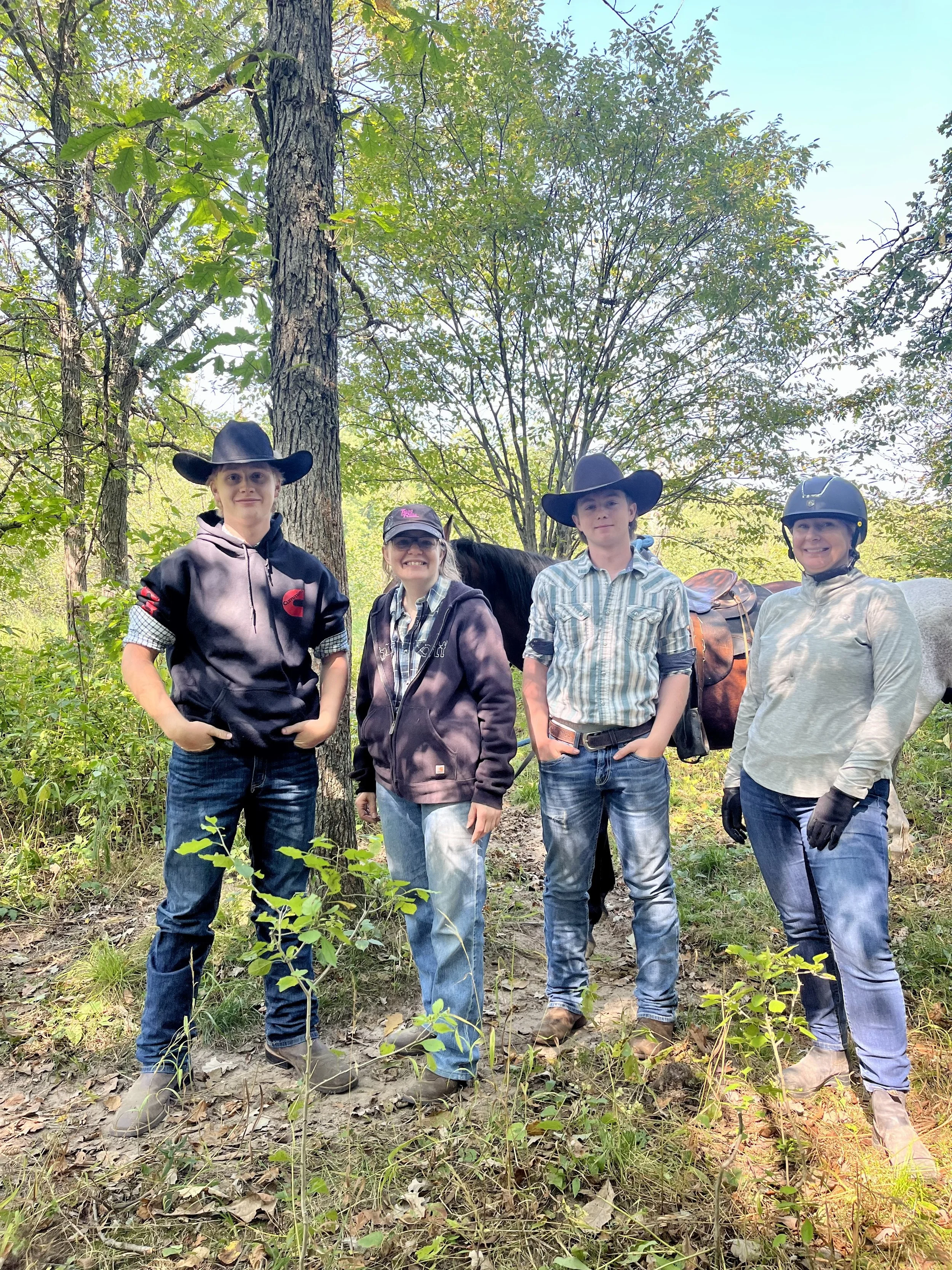 Four people standing in a wooded area, wearing casual outdoor clothing and hats, with a horse in the background.