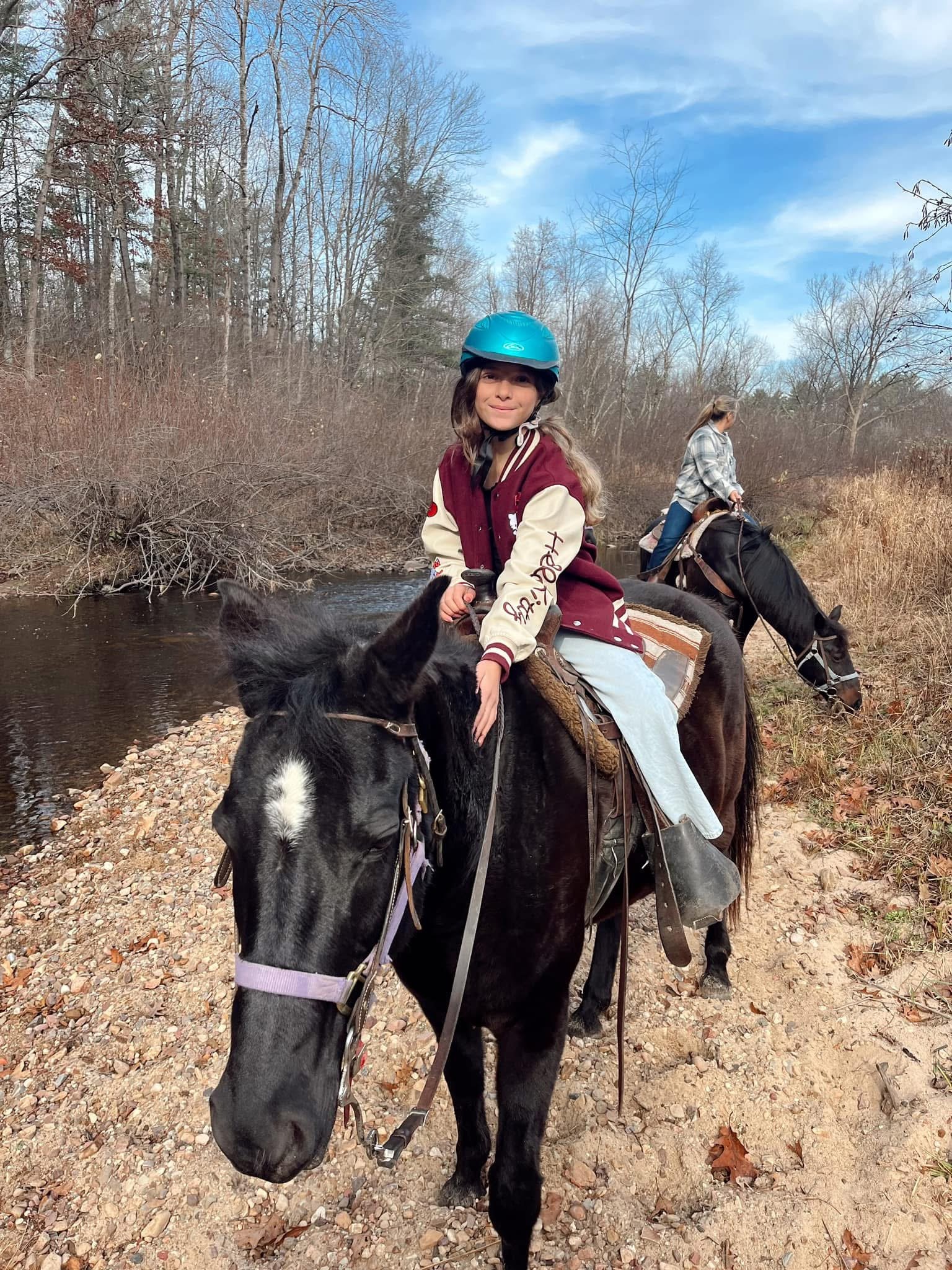 A young girl wearing a blue helmet is riding a black horse through a wooded area with a small stream and leafless trees, accompanied by an adult woman on horseback in the background.