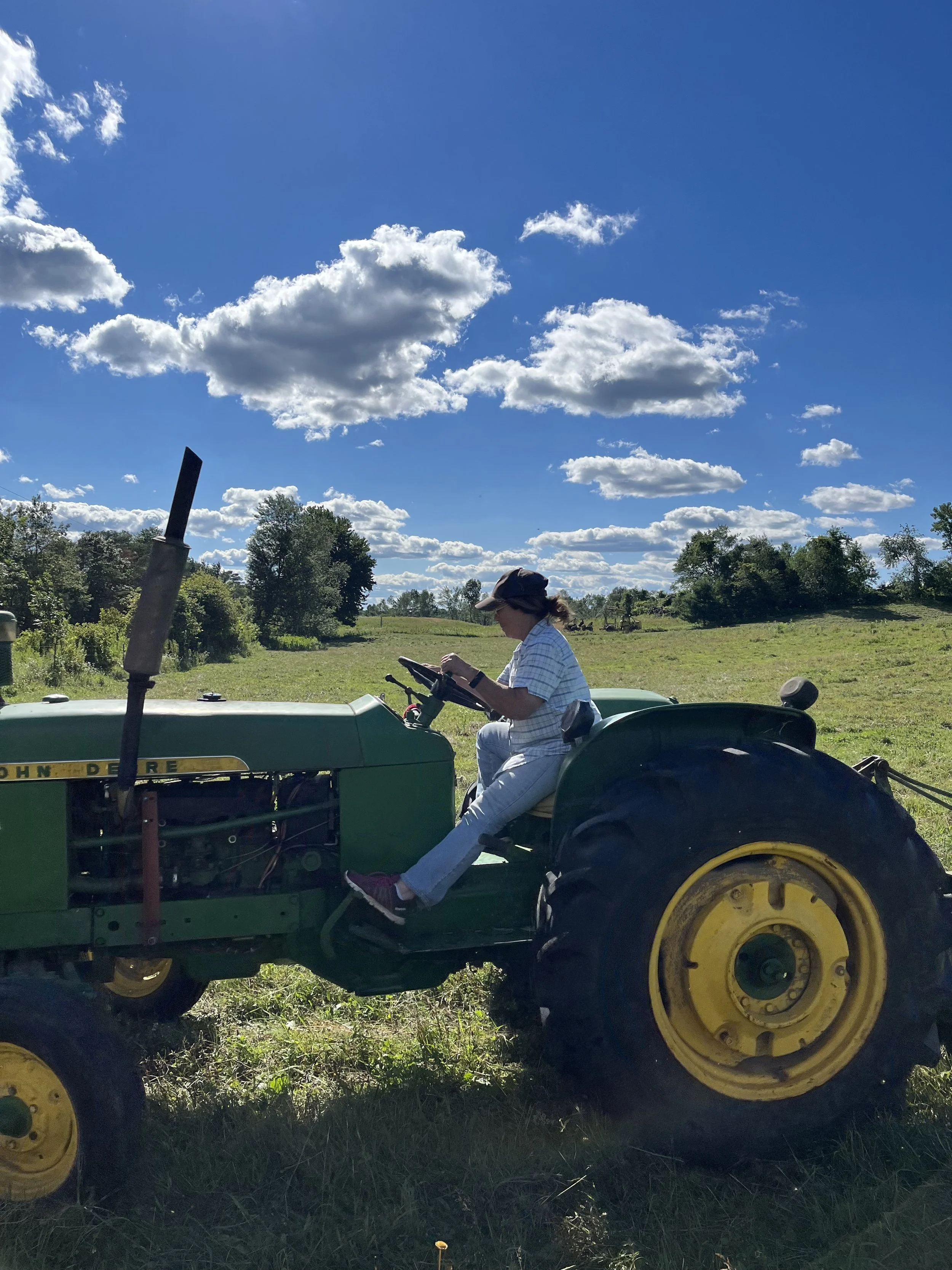 Person sitting on a green John Deere tractor in a grassy field under a partly cloudy blue sky, wearing a hat and using a tablet.
