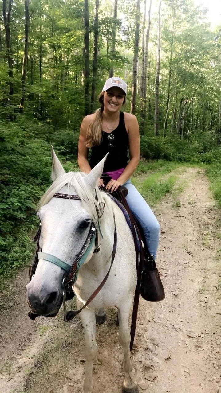 A young woman riding a white horse on a dirt trail in a wooded area with green trees and sunlight filtering through.
