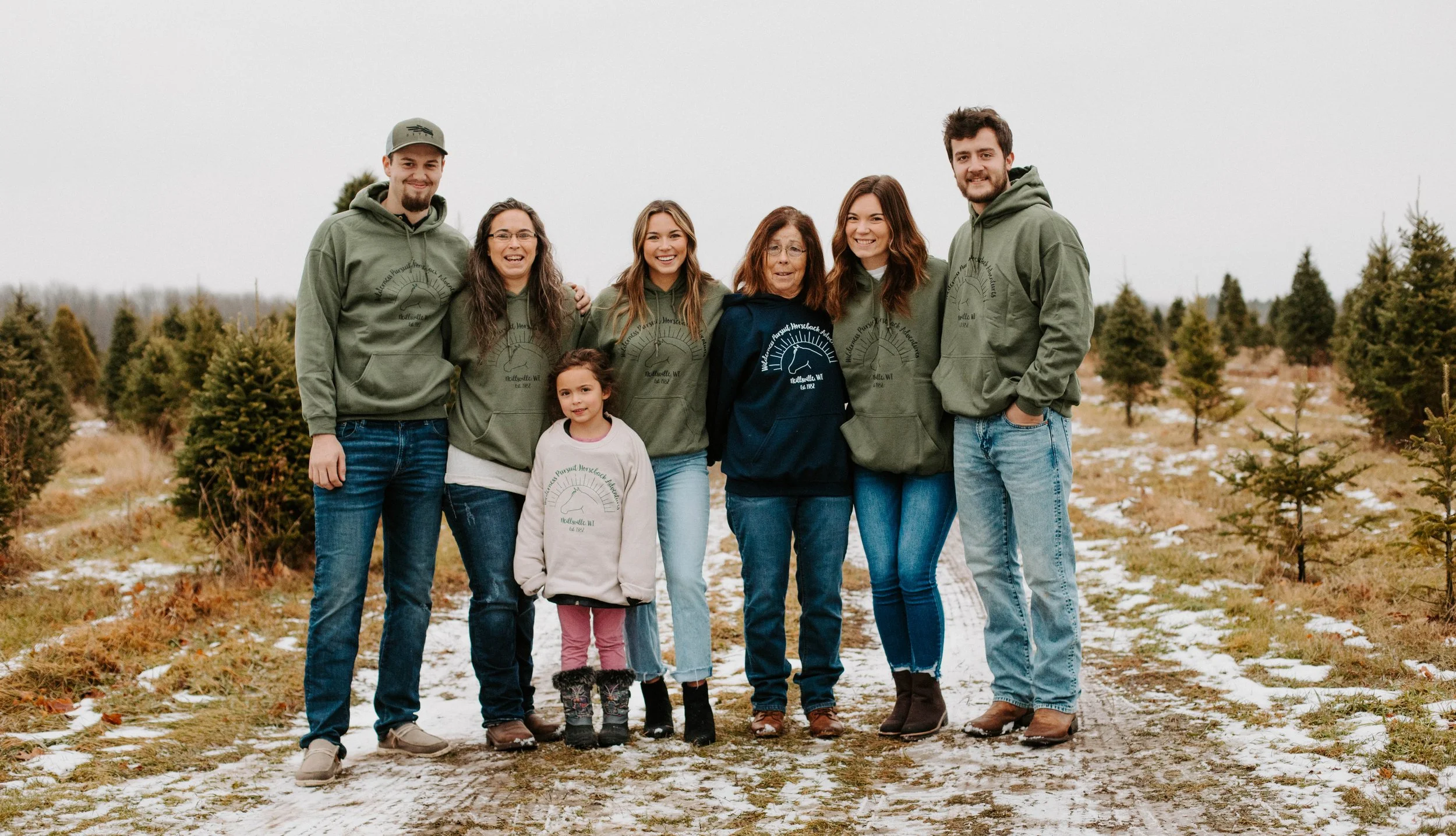 A group of seven people, including children and adults, standing outdoors on a dirt path surrounded by small evergreen trees, with some patches of snow on the ground. They are dressed in casual clothing, with some wearing matching green hoodies with a logo.