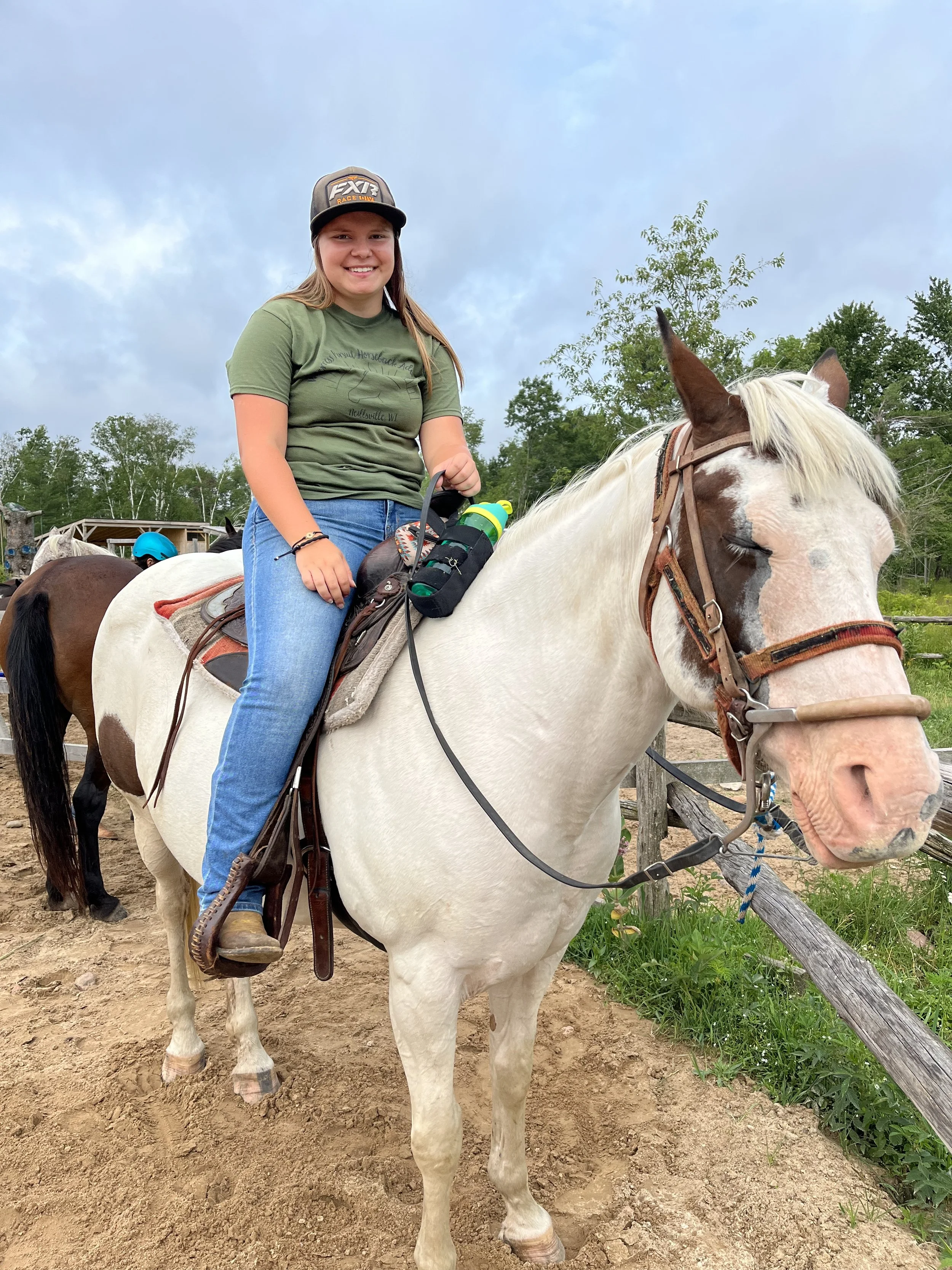 A young girl with long hair, smiling, wearing a baseball cap, a green t-shirt, and jeans, riding a white horse with brown patches, in an outdoor setting with trees and cloudy sky.