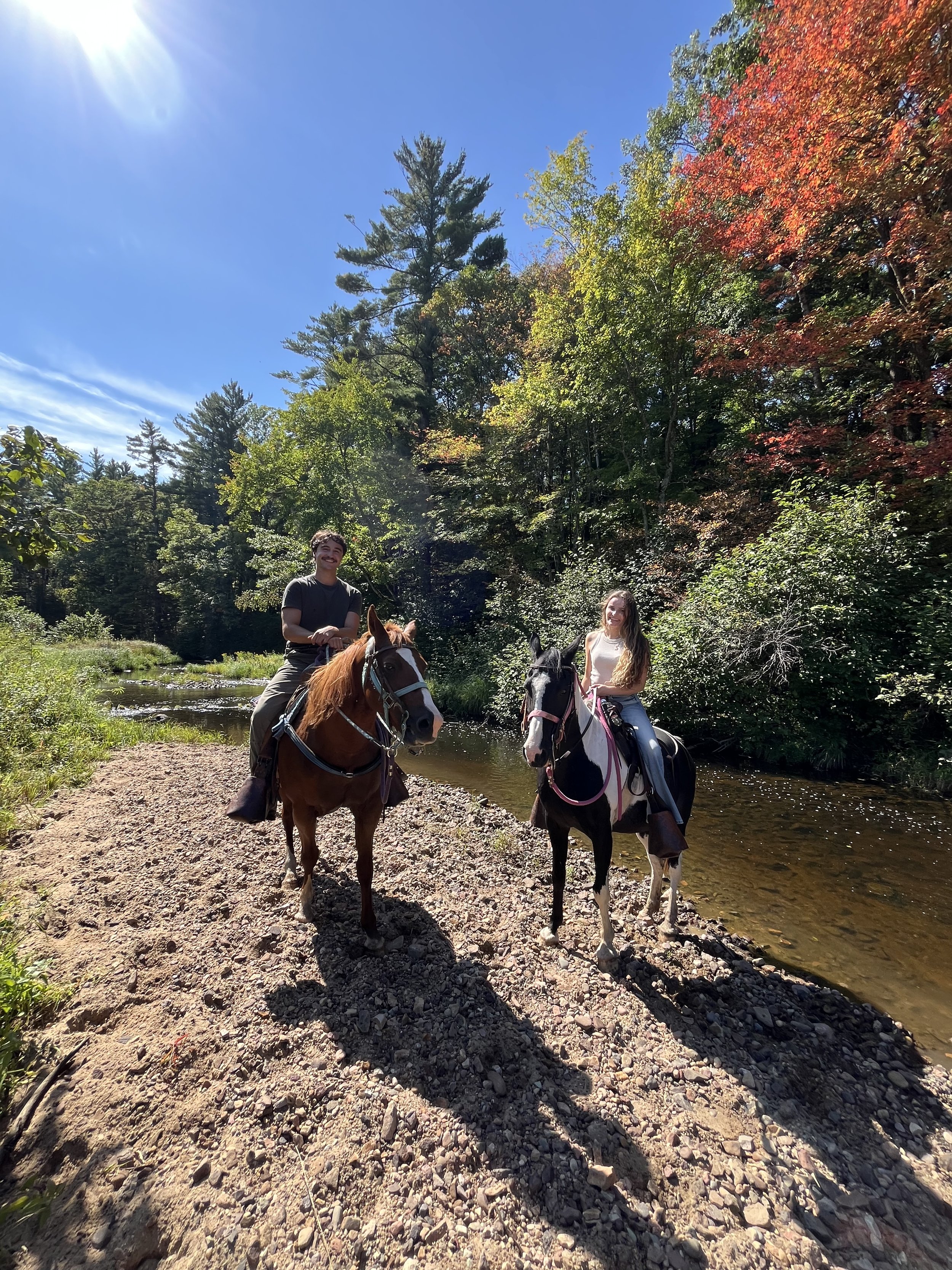 Two people riding horses by a creek in a wooded area during fall with colorful leaves and a clear blue sky.