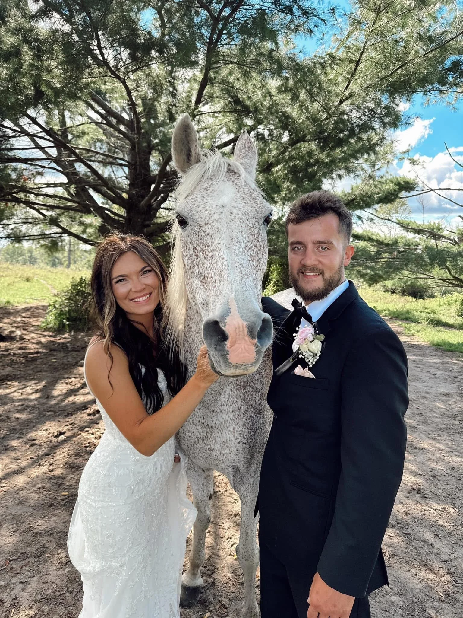 A couple in wedding attire embracing a gray horse outdoors with trees and a blue sky in the background.