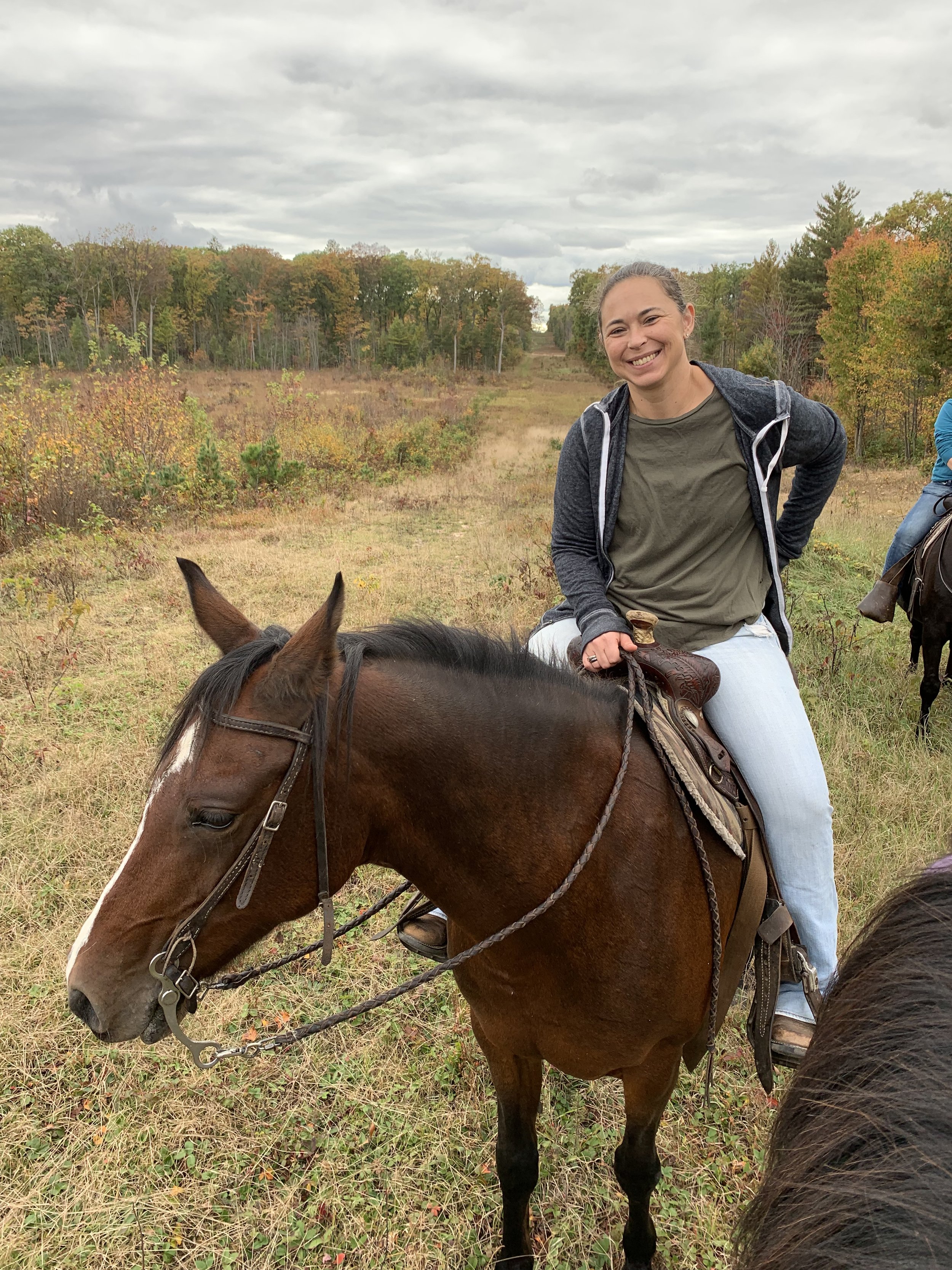 A woman riding a horse on a grassy trail in a wooded area during autumn, with clouds overhead.