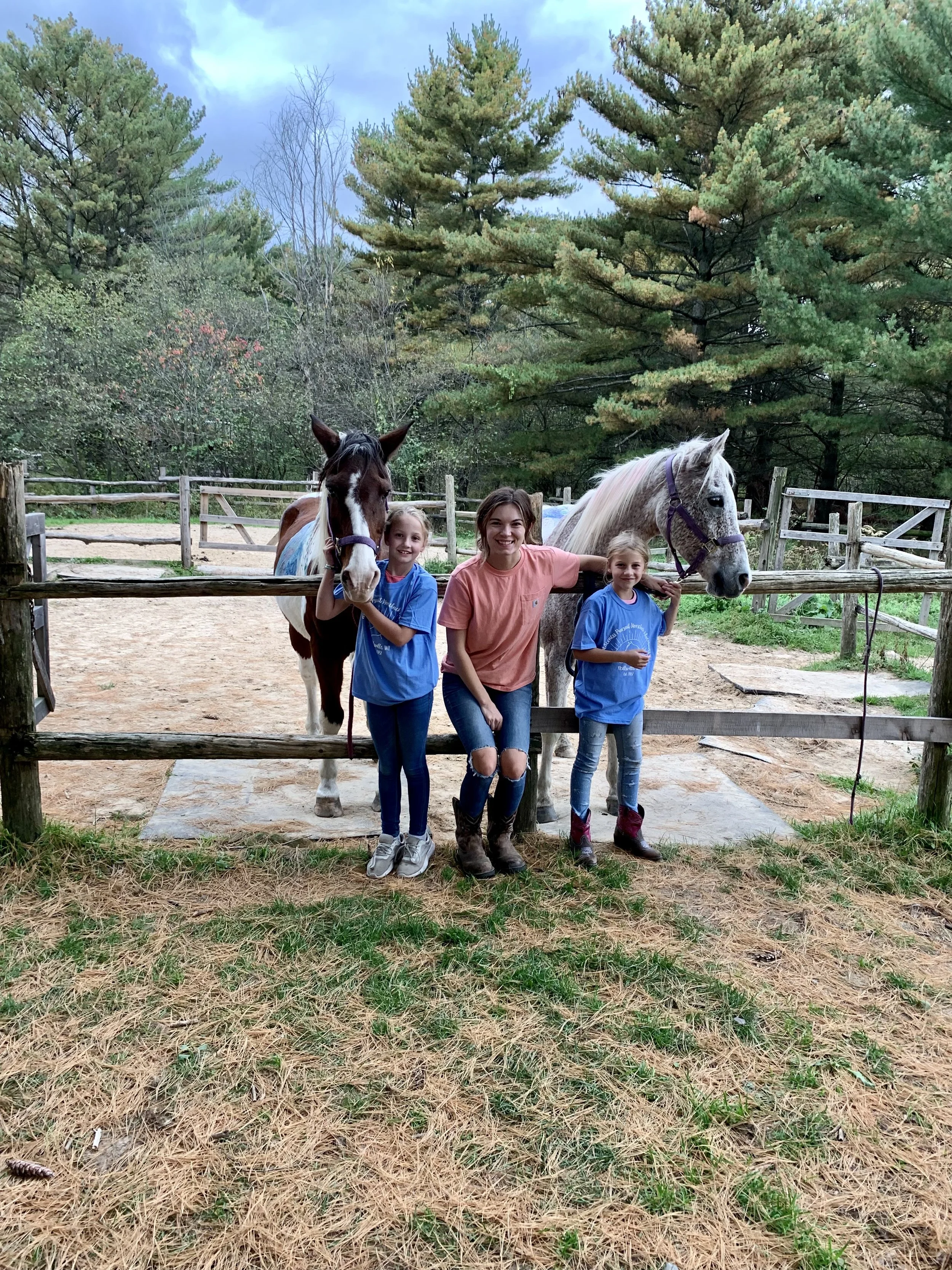 Three children and an adult standing between two horses in a fenced outdoor area with tall pine trees and a cloudy sky in the background.