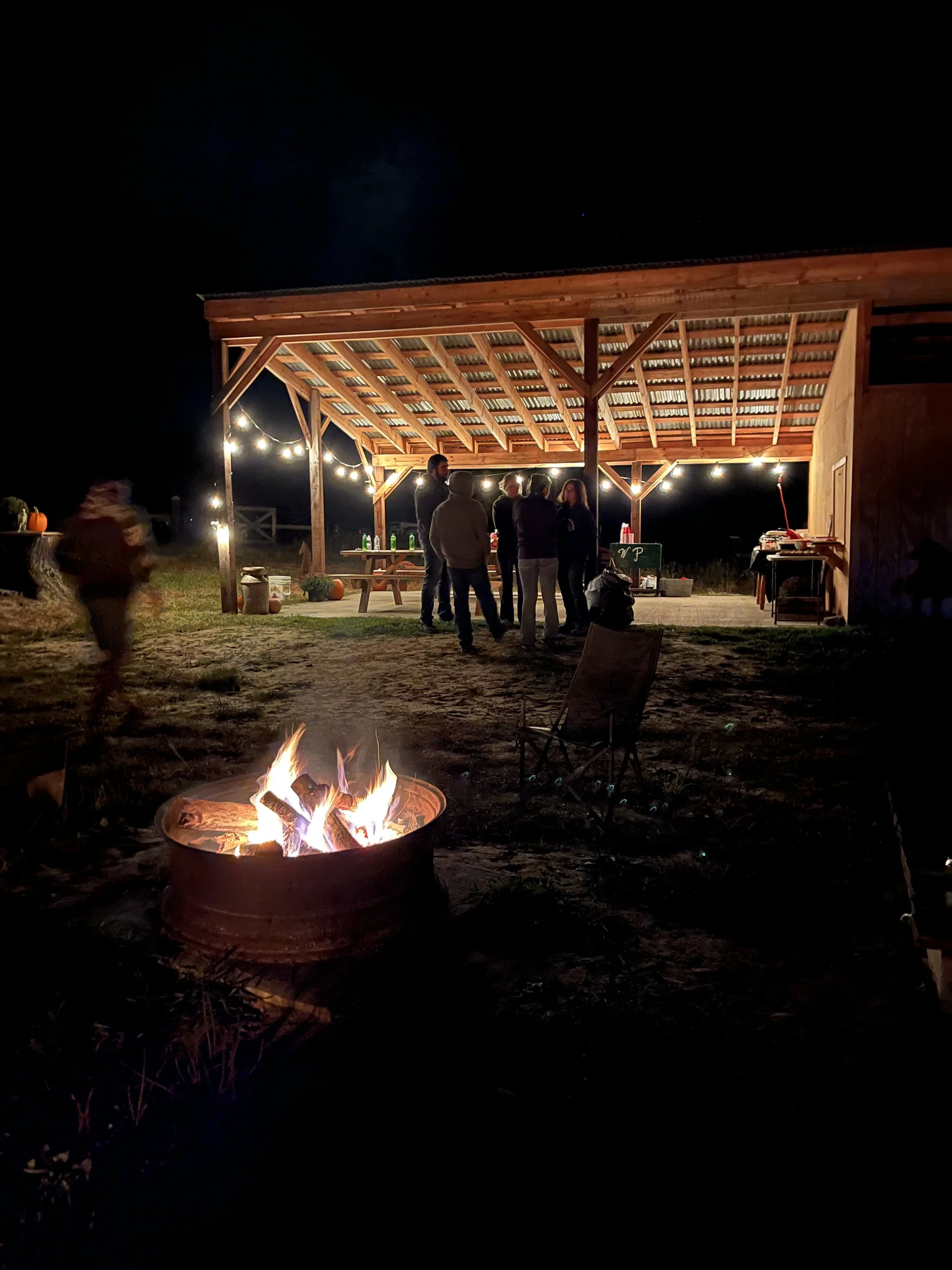 Night scene of a group of people gathered under a wooden shelter with string lights, a fire pit with a burning fire in the foreground, and pumpkins on the left side.