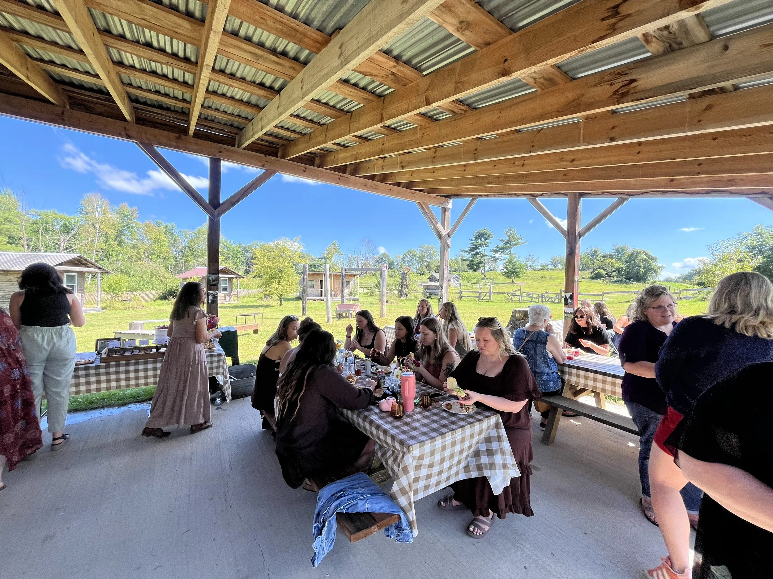 People sitting at picnic tables under a rustic wooden pavilion on a sunny day, with a grassy hill and small buildings in the background.