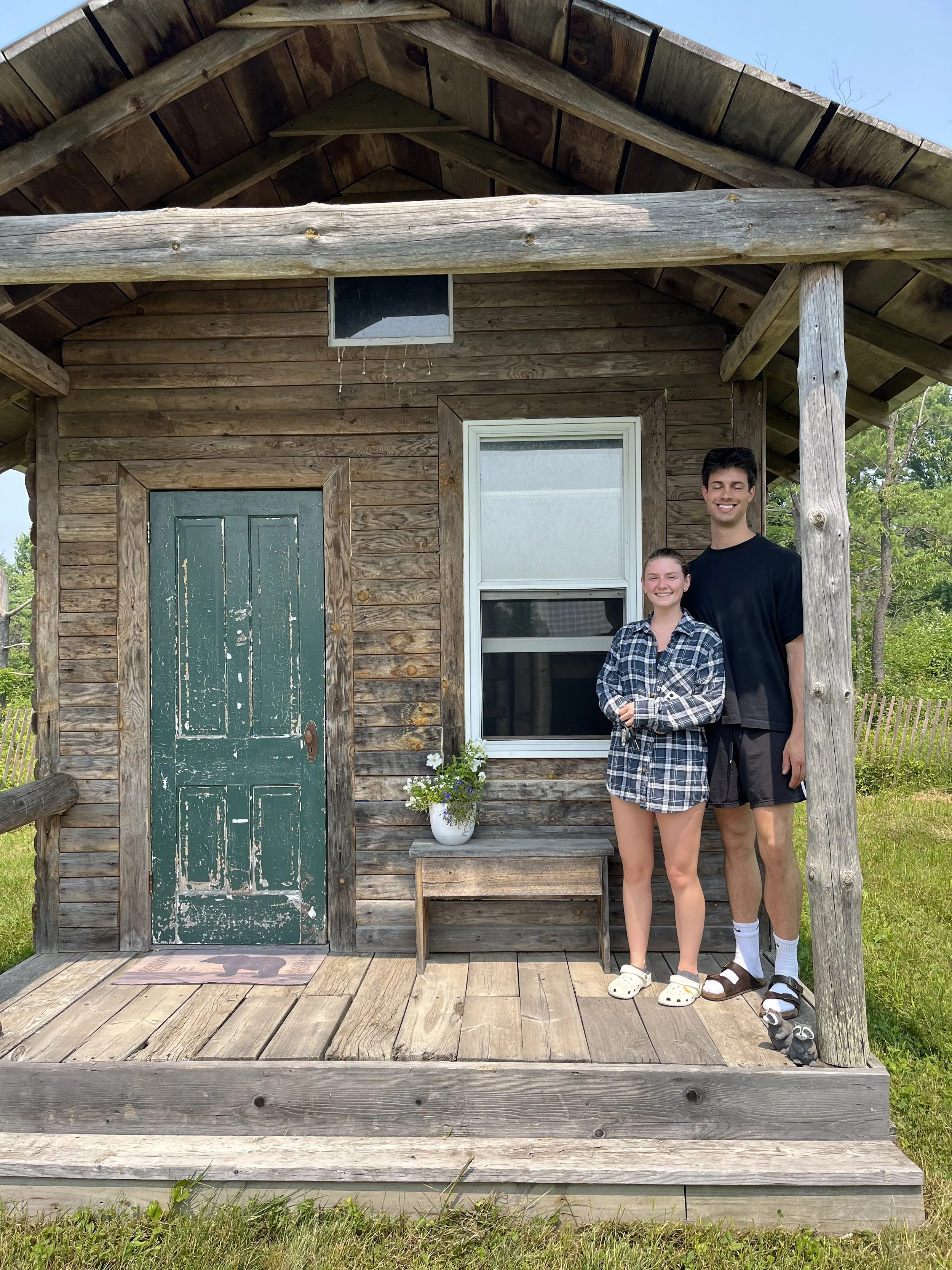 A young couple standing on the porch of a small wooden cabin, smiling at the camera. The cabin has a green weathered door, a window, and a small table with a potted plant outside. The porch is made of wooden planks, and the background shows trees and greenery.