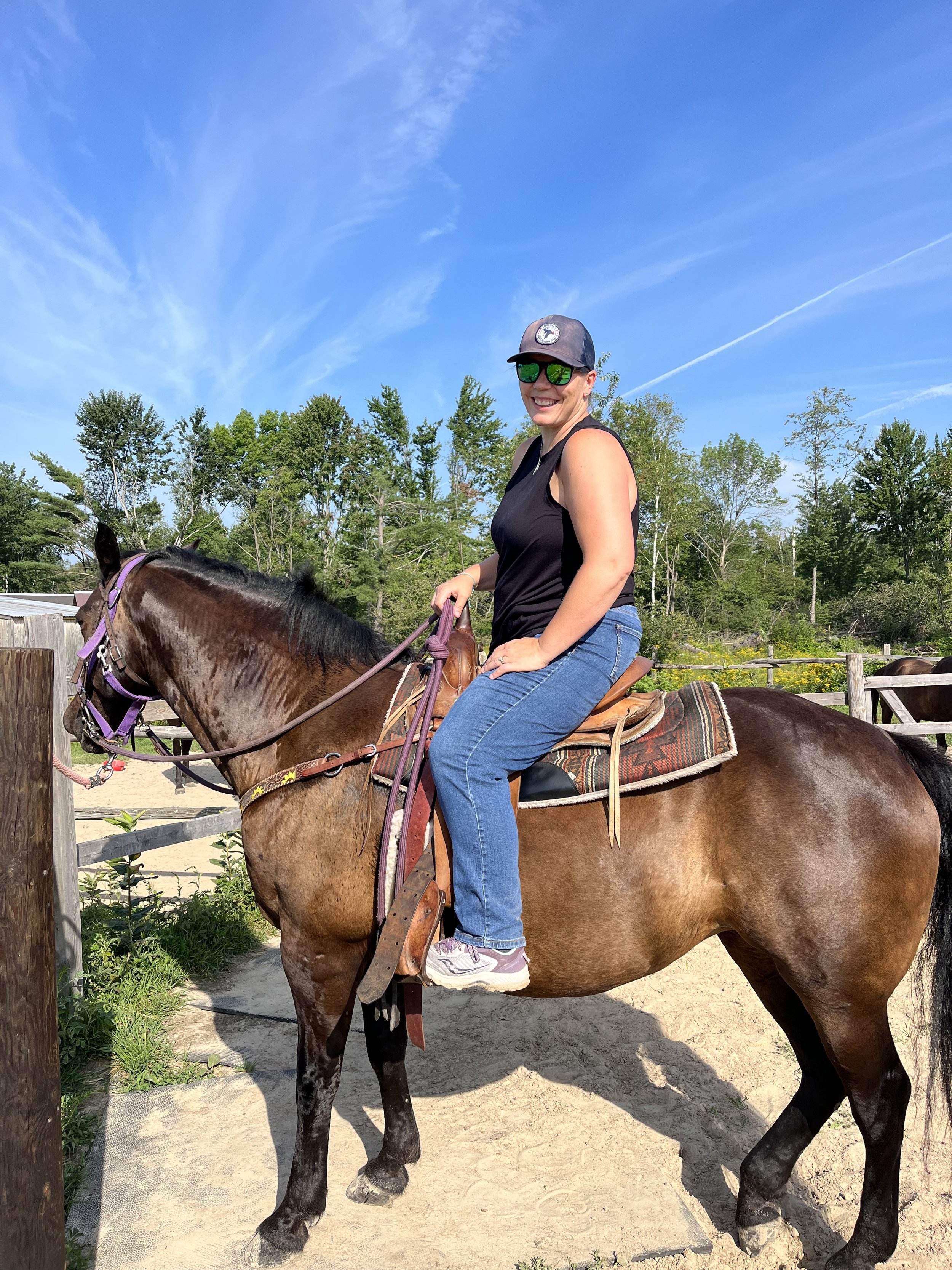 A woman riding a brown horse outdoors under a blue sky with a few clouds, wearing sunglasses, a cap, a black tank top, and jeans.