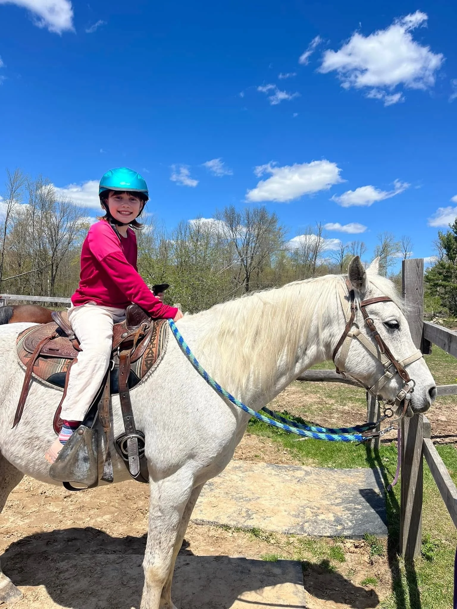 A young girl wearing a pink jacket and blue helmet rides a white horse with a saddle and bridle, outdoors on a sunny day under a blue sky with some clouds.