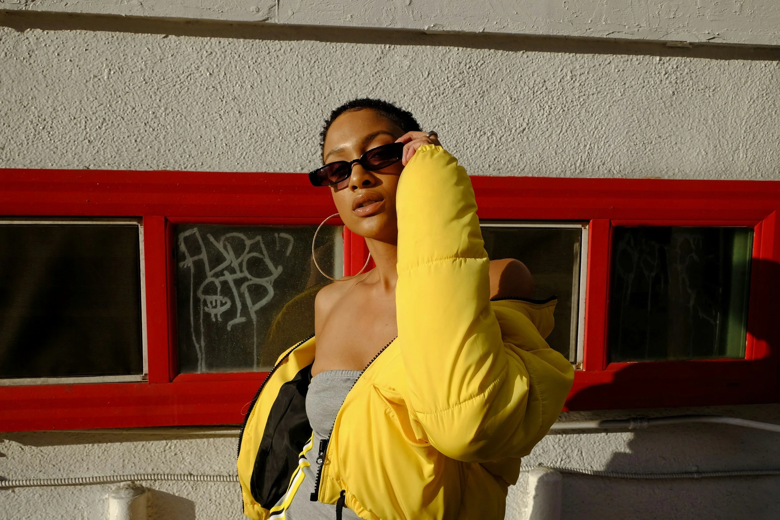 A woman wearing sunglasses and a yellow jacket standing in front of a red-framed window with graffiti, casting a shadow on a white wall.