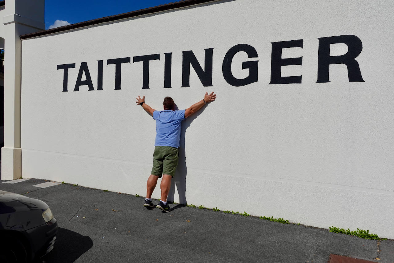 En person står framför en vit vägg med stora svarta bokstäver som stavar 'TAITINGER' och sträcker ut armarna.