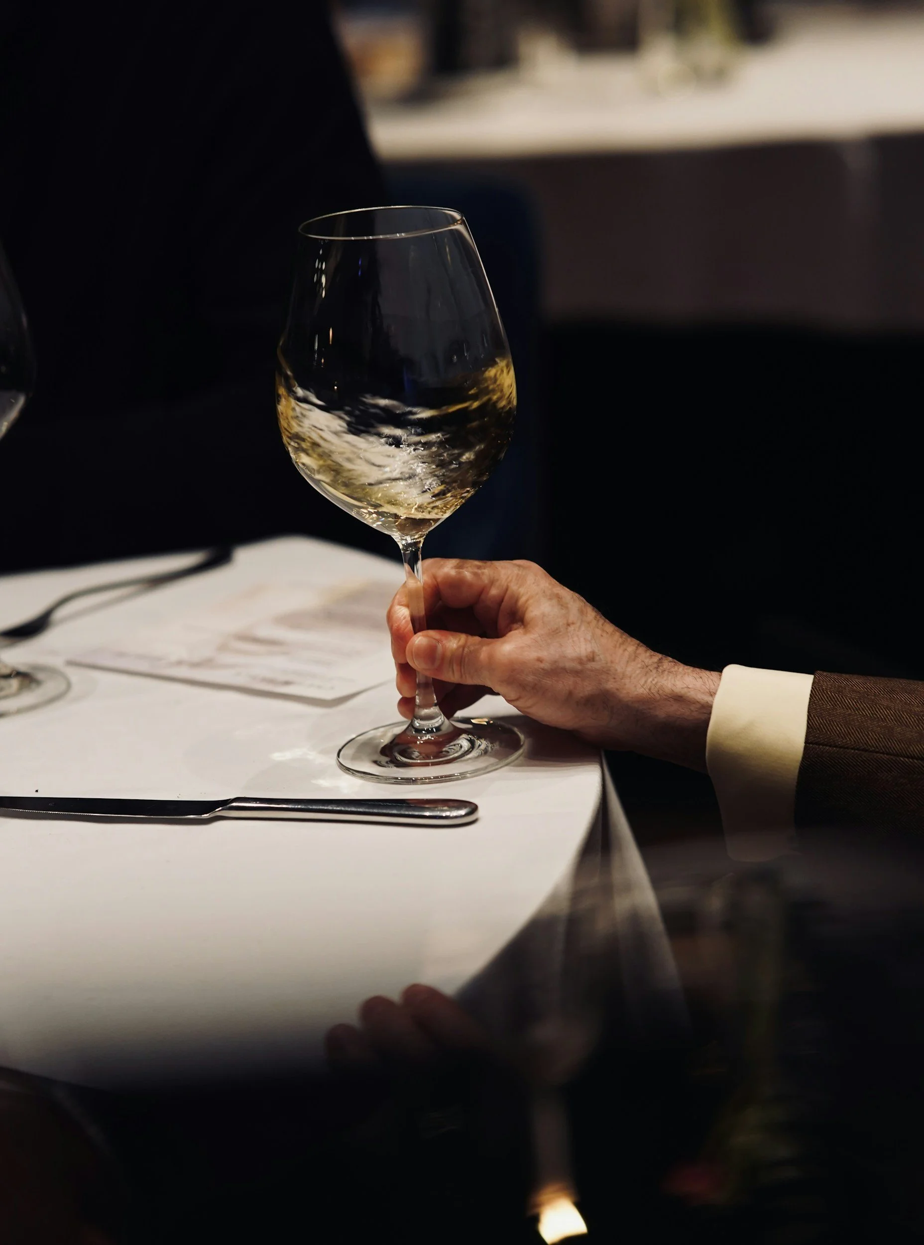 En person håller ett glas vitt vin på en restaurangbord, med bestick och en meny synliga.