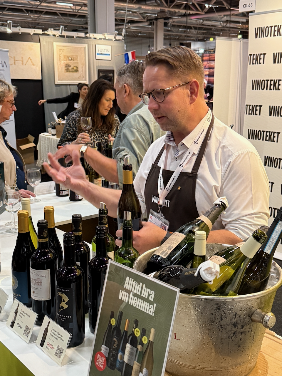 Man som serverar vin vid vinmässa, omgiven av vinflaskor och en kylväska med svalkade flaskor.